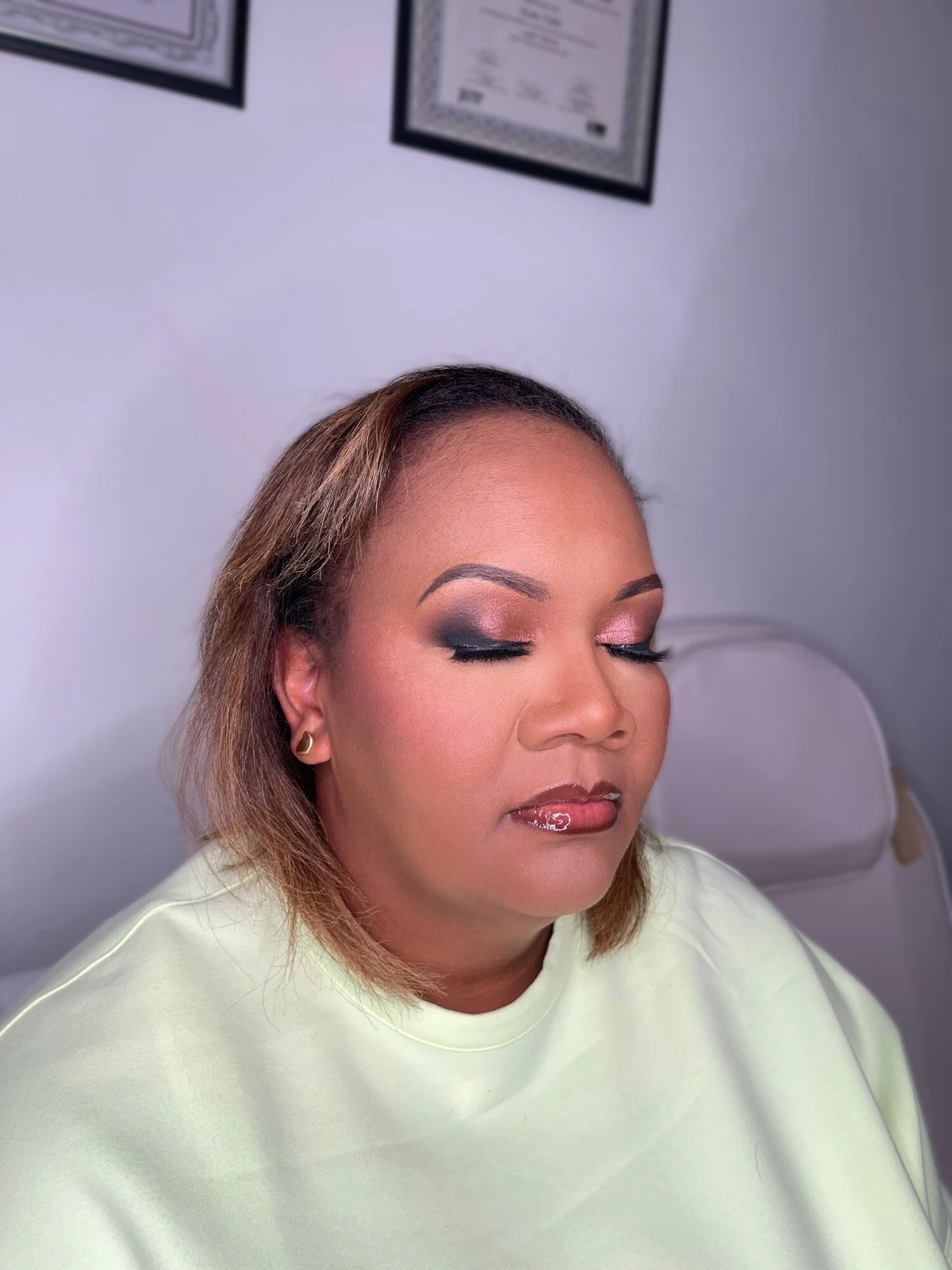 A woman with makeup, earrings, and a light yellow top, resting at a medical or beauty appointment, with framed certificates on the wall behind her.
