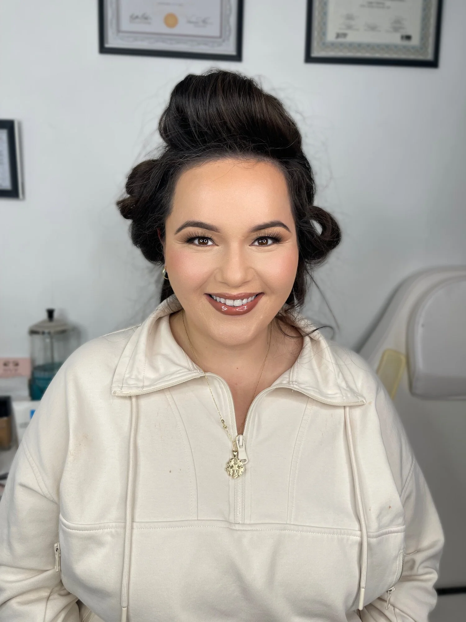 A woman with dark, styled hair smiling in a white zip-up jacket, with certificates hanging on the wall behind her.