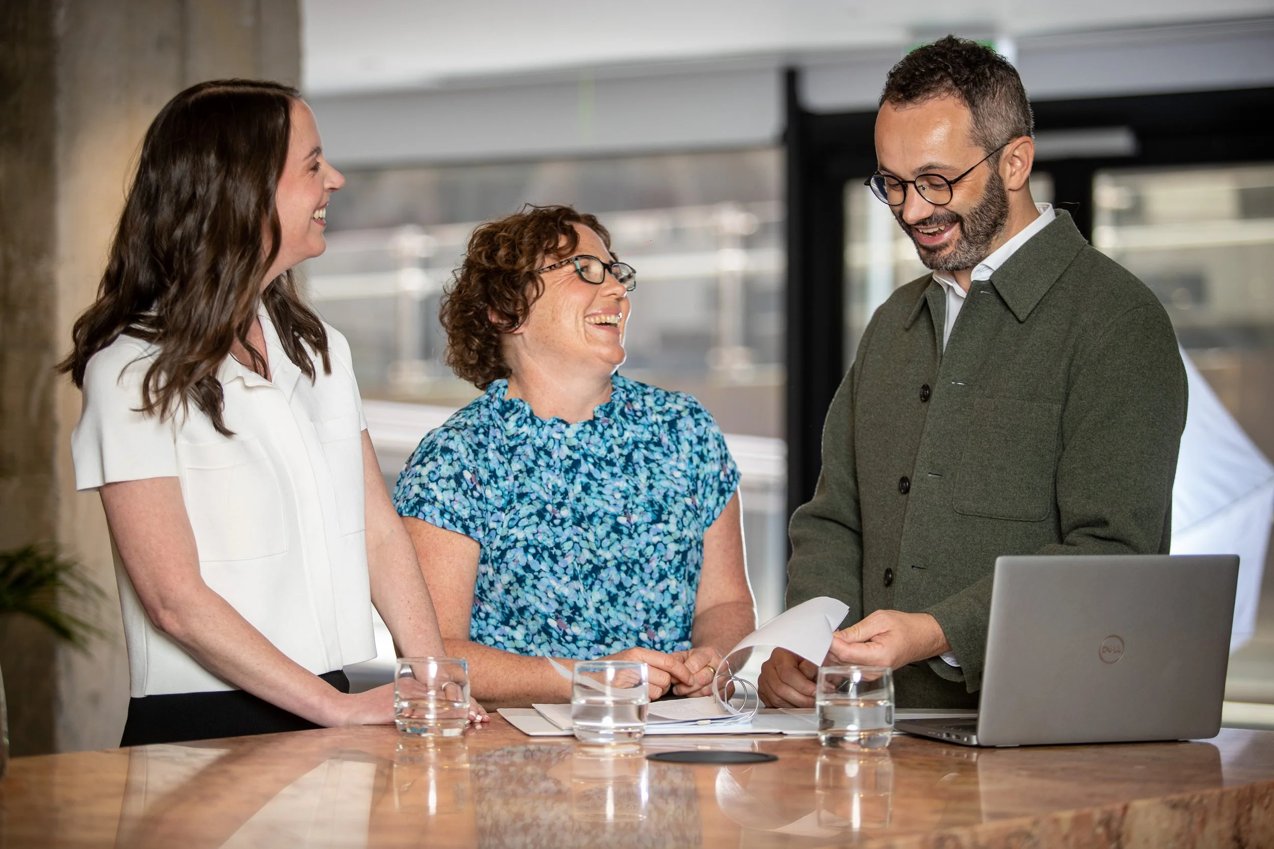 Three people, two women and one man, are smiling and engaging in conversation in an office setting with a laptop and documents on the table.