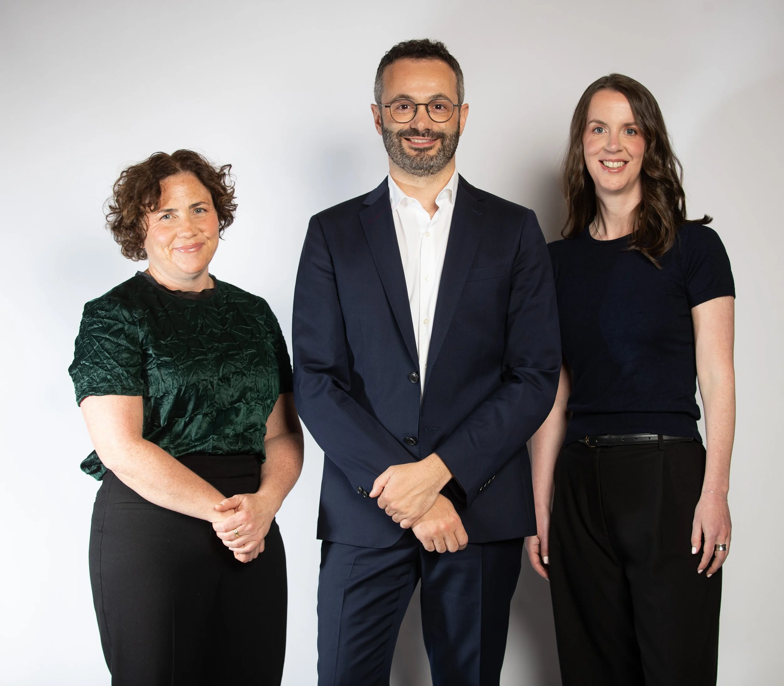Three professionals standing together on a white background, smiling at the camera. From left to right, a woman with curly brown hair wearing a green textured blouse and black pants, a man with glasses and a beard in a dark suit with a white shirt, and a woman with long brown hair in a black top and black pants.