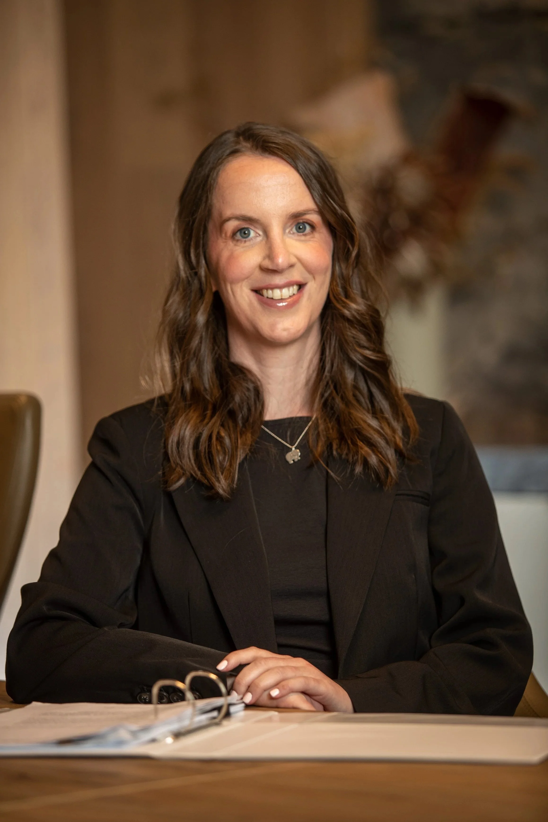 A woman with long brown hair and blue eyes, wearing a black blazer and a necklace with an elephant pendant, smiling while sitting at a table with a binder and papers.
