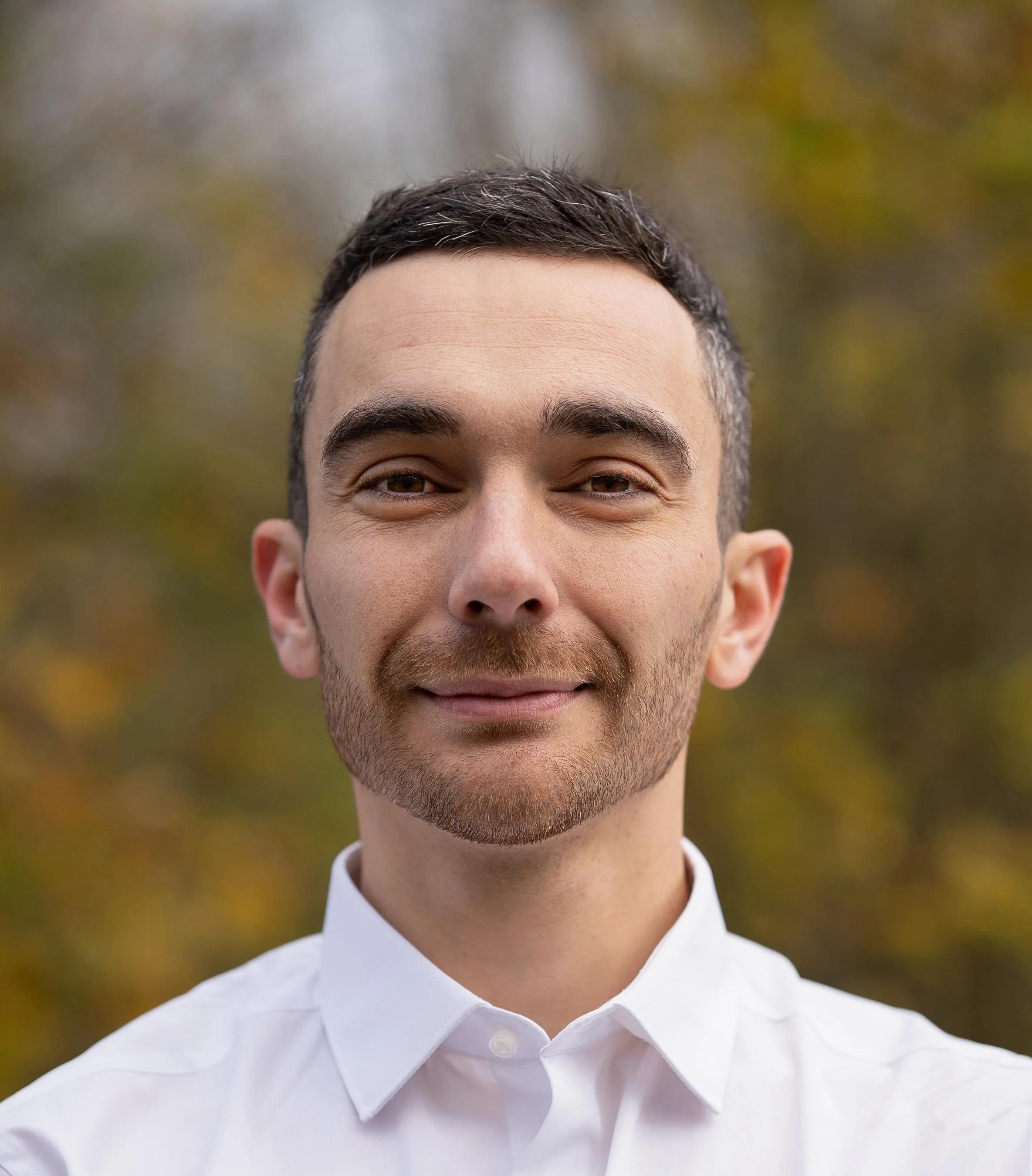 A smiling man with short dark hair, facial hair, wearing a white collared shirt, outdoors with blurred fall foliage background.