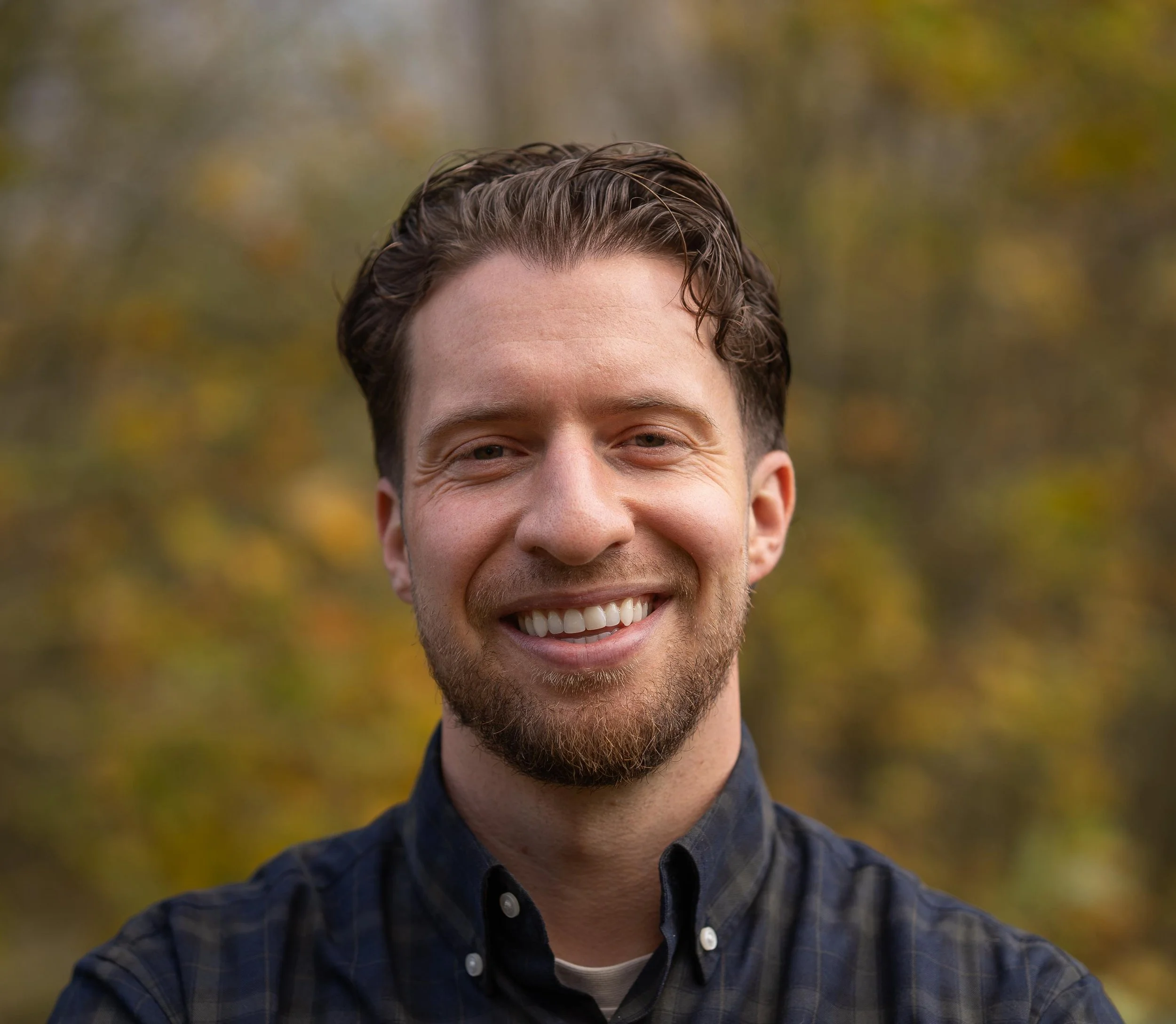 A smiling man with short brown hair and a beard, wearing a dark plaid shirt, outdoors with autumn-colored trees in the background.