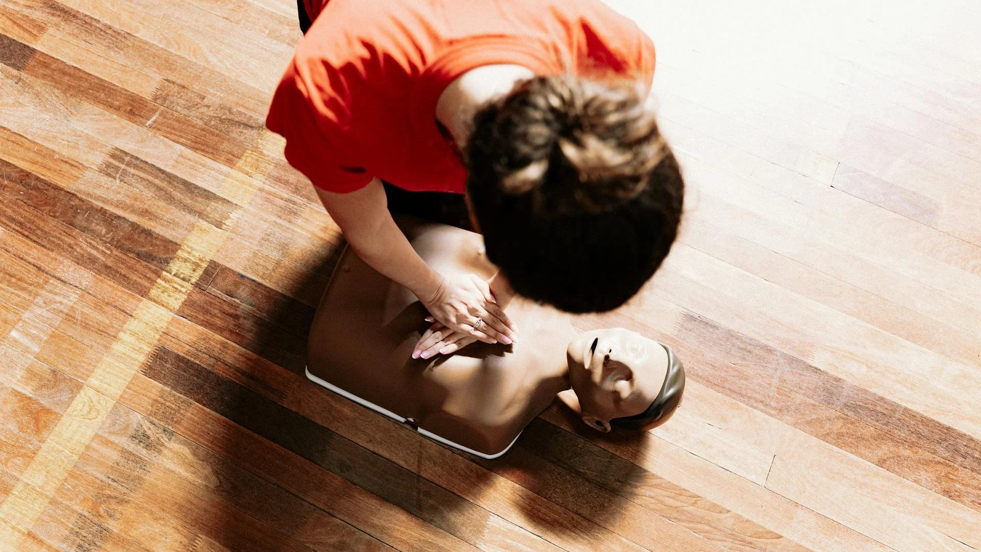Person performing CPR on a training mannequin on a wooden floor.