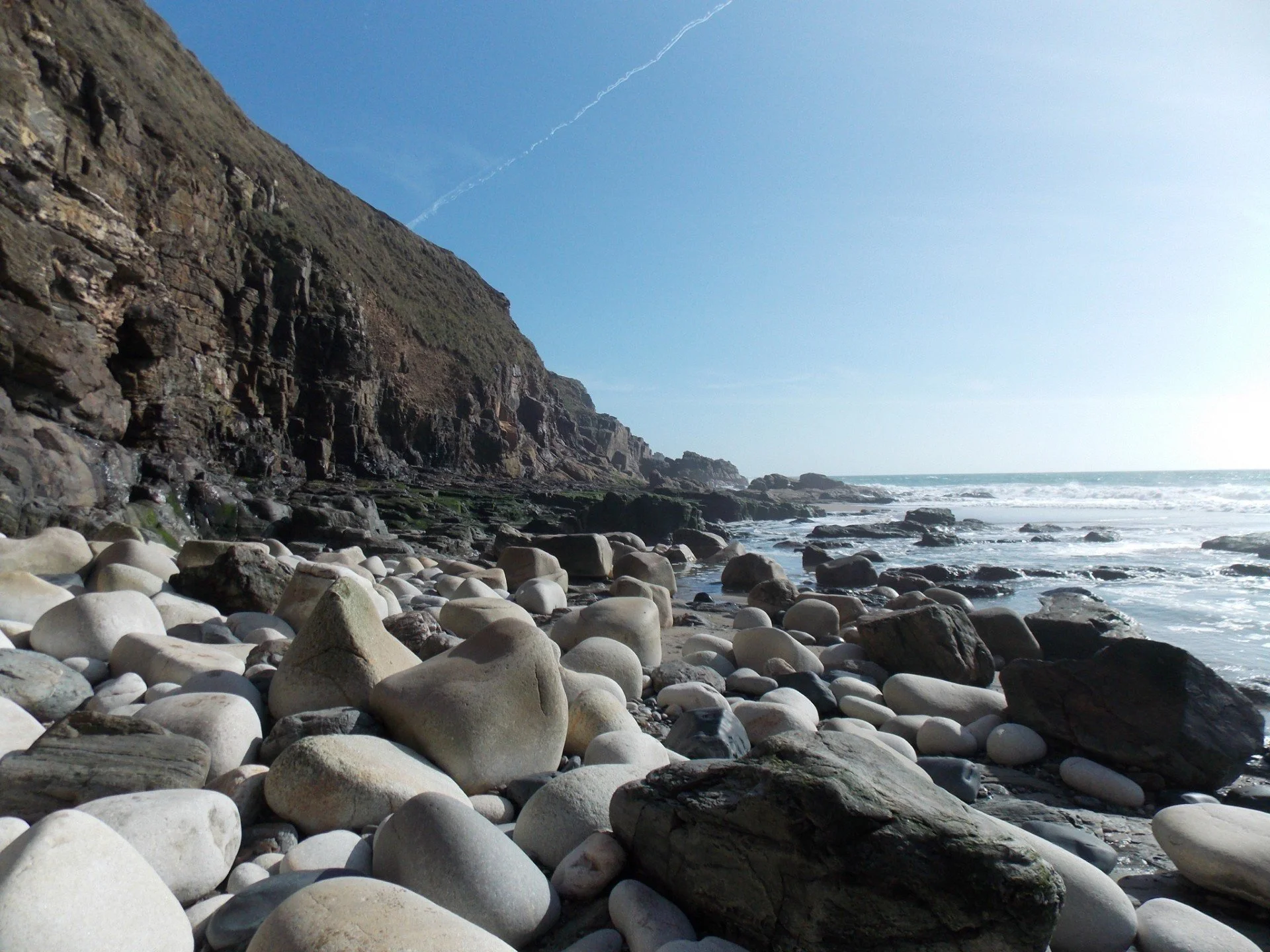 Rocky shoreline with large, smooth stones in the foreground, a steep cliff on the left, and waves crashing onto the beach under a clear blue sky.