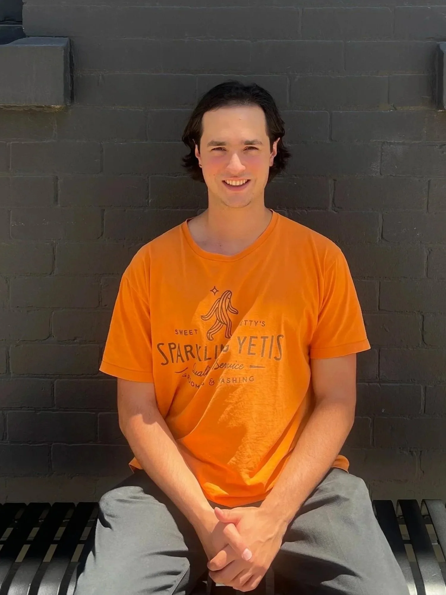 A young man with dark hair, wearing an orange t-shirt and light-colored pants, is sitting on a bench against a dark brick wall, smiling at the camera.