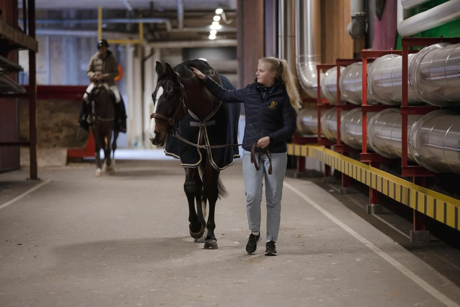 A woman petting a black horse with a white marking on its face inside an indoor stable. There is another person on horseback in the background, and large metal pipes are stored on the right side of the scene.