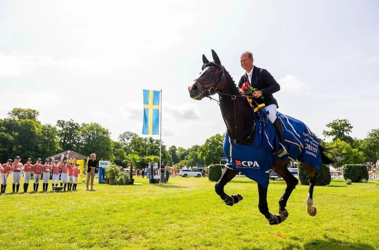 A man riding a horse in a race or competition, holding flowers, with a Swedish flag in the background and a team of people in historical costumes watching.