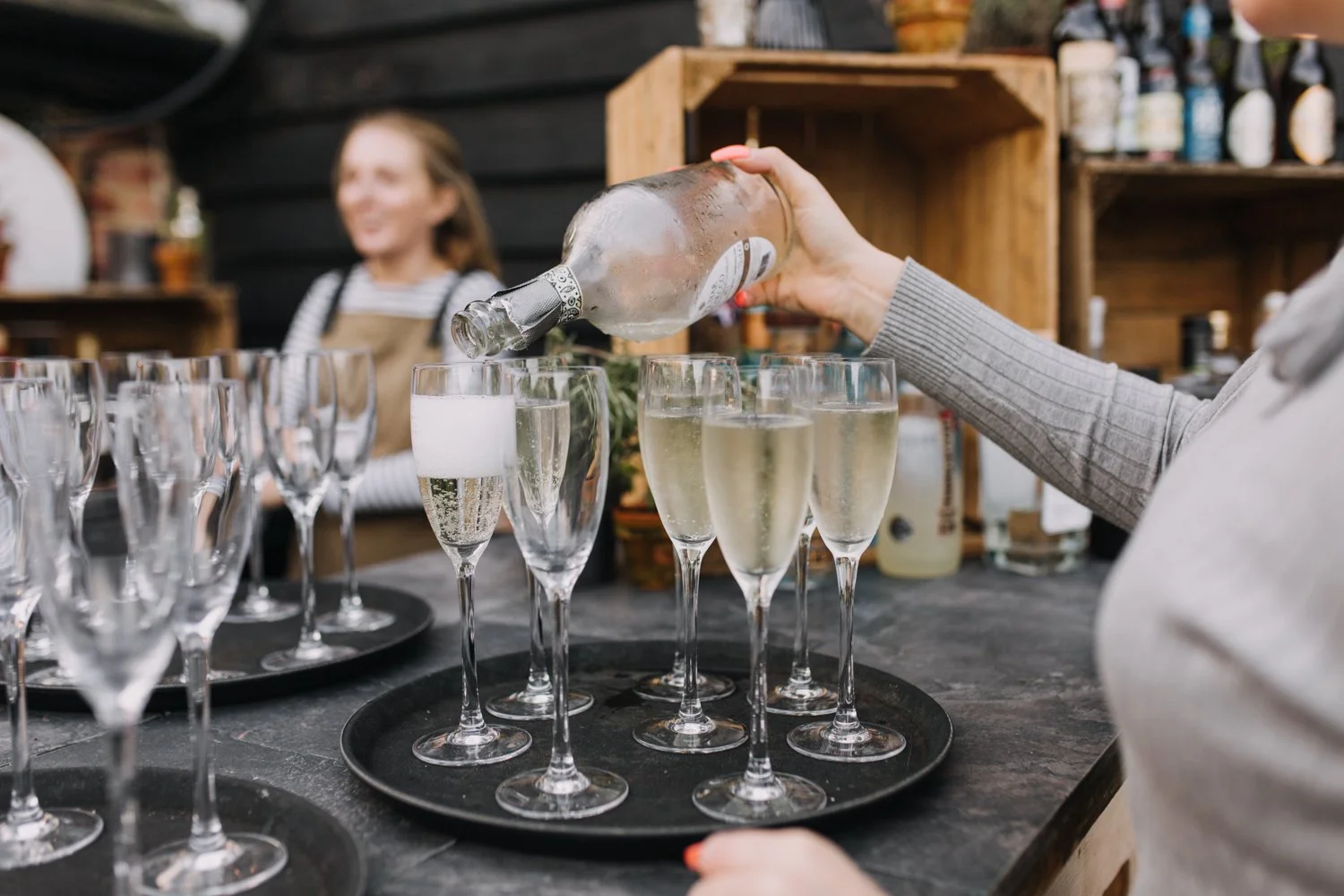 Person pouring champagne into glasses at a celebration or event, with a woman smiling in the background.
