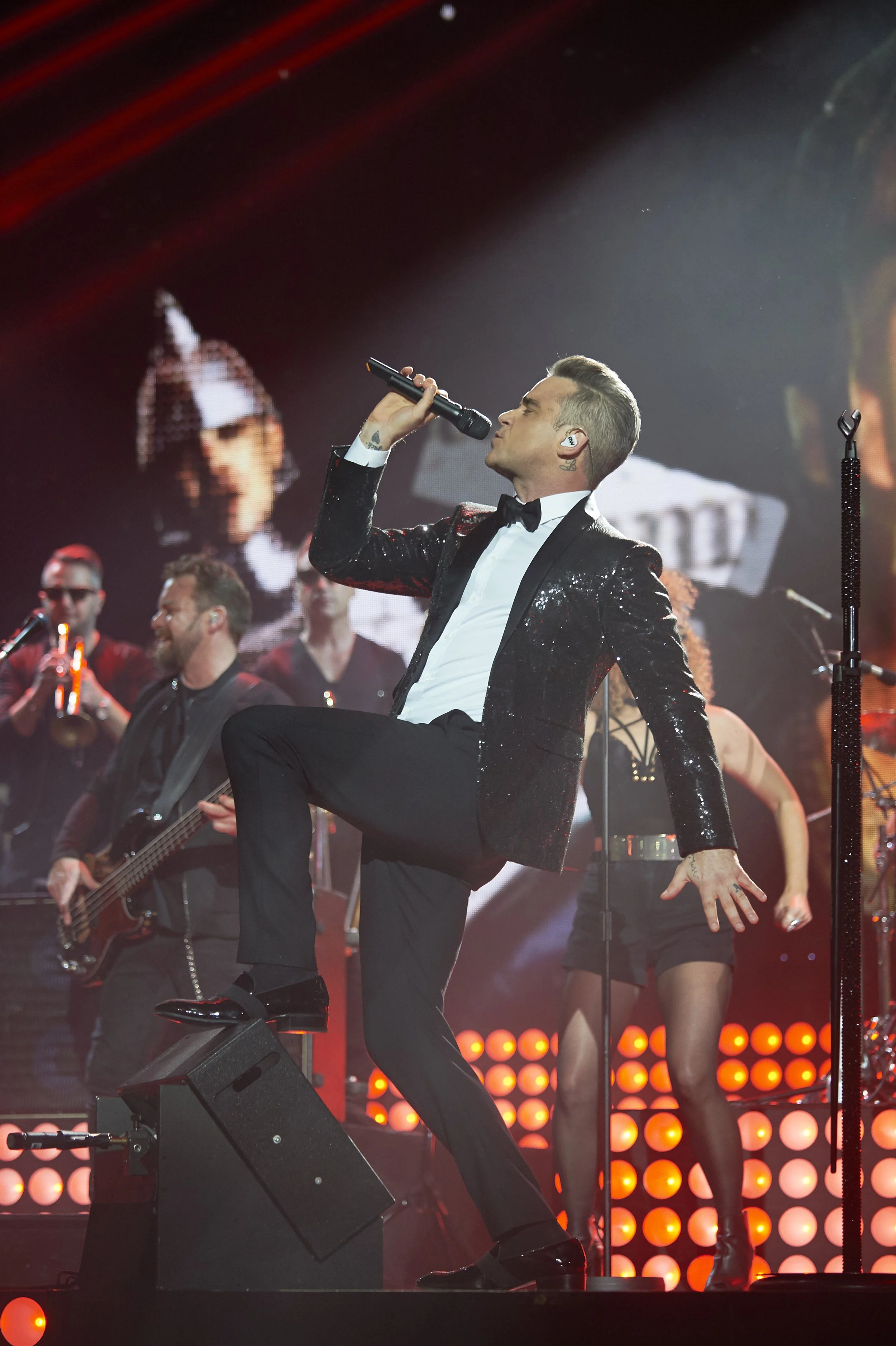 A male singer in a black sequined tuxedo jacket, white shirt, and black pants performing on stage with a microphone. Other musicians and backup singers are visible in the background, and the stage is lit with red and orange lights.