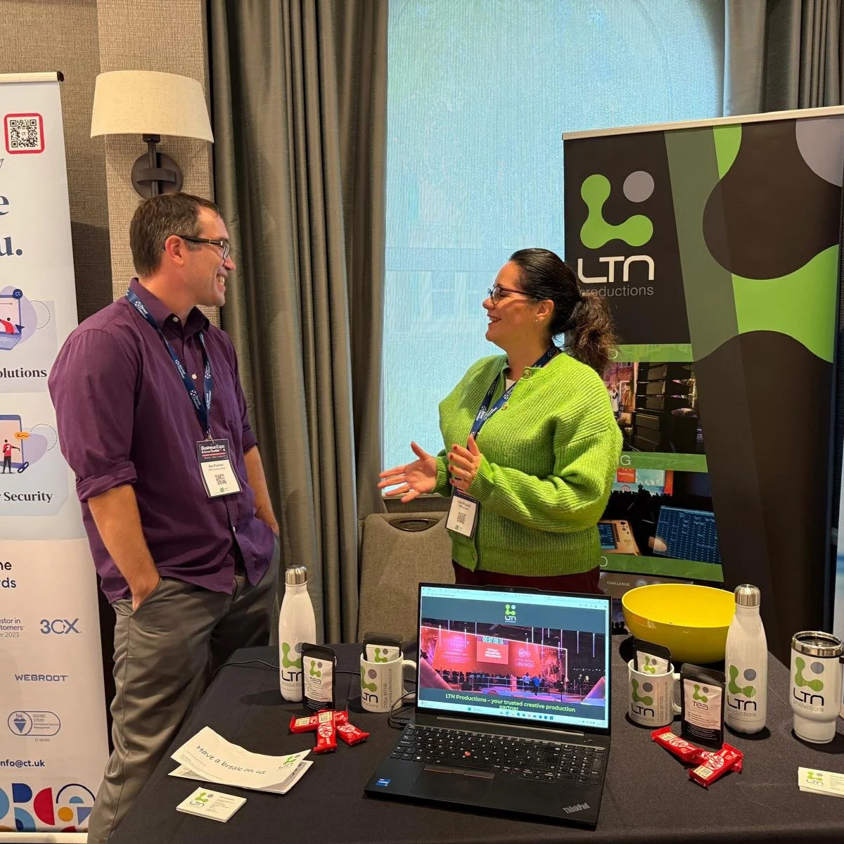 Two people talking at a booth with promotional materials and a laptop on a table, with banners displaying the LTN Productions logo.