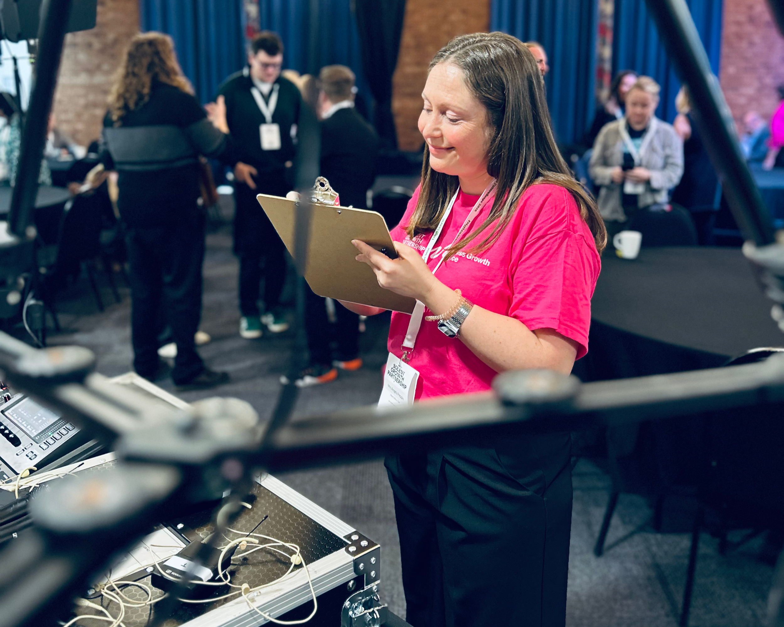 A woman in a pink shirt holding a clipboard, standing in a busy conference room with people in the background, some engaged in conversations, and conference equipment in the foreground.