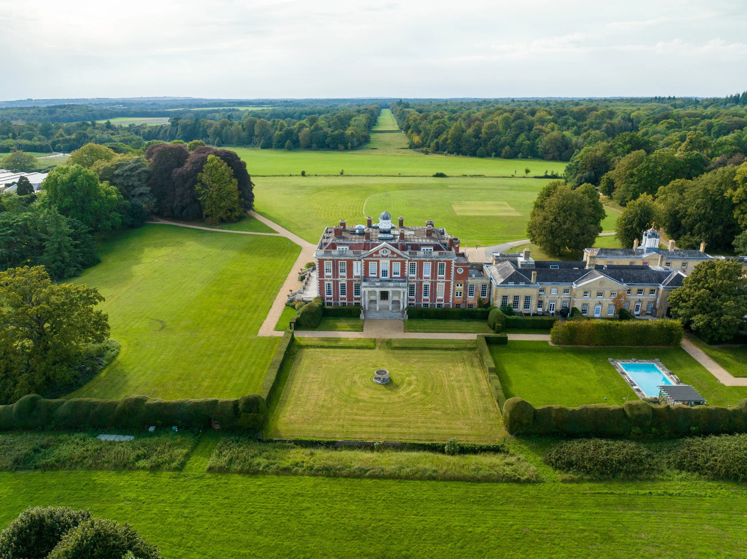 Aerial view of a large, historic estate with a red brick main building, surrounded by neatly maintained lawns, gardens, a swimming pool, and expansive green fields and trees in the background.