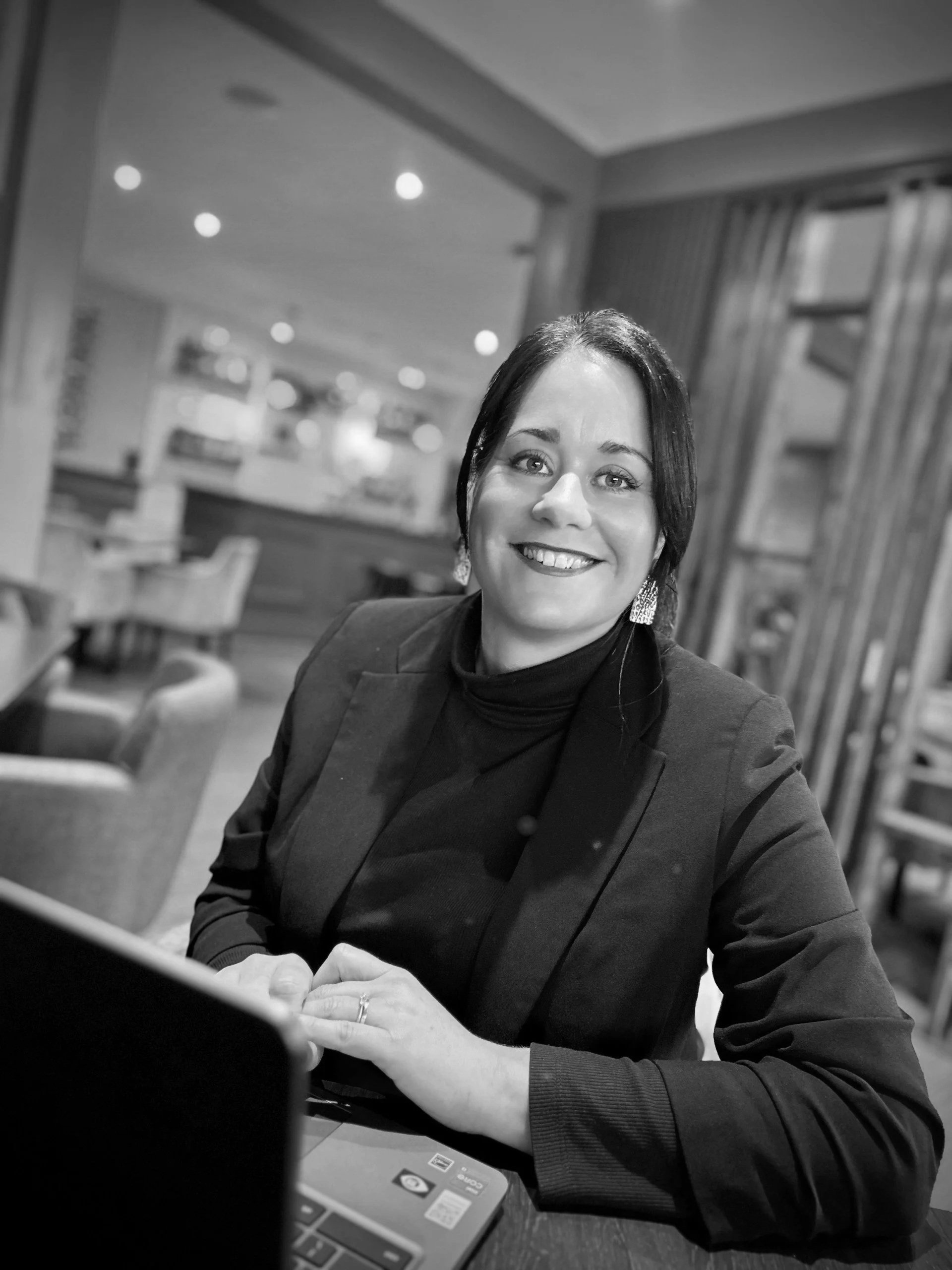 A woman with dark hair smiling, wearing a dark blazer and earrings, sitting at a table in a restaurant or cafe, with a laptop in front of her.