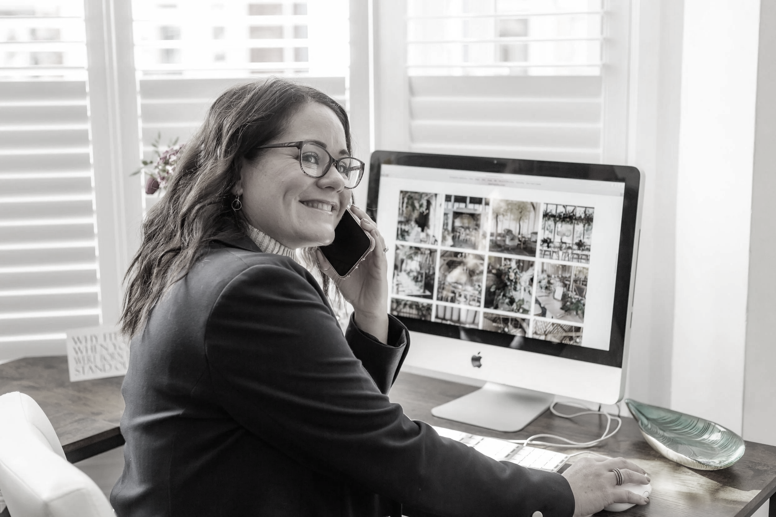 A woman with glasses smiling while talking on a phone at her desk with an iMac computer displaying a photo gallery.