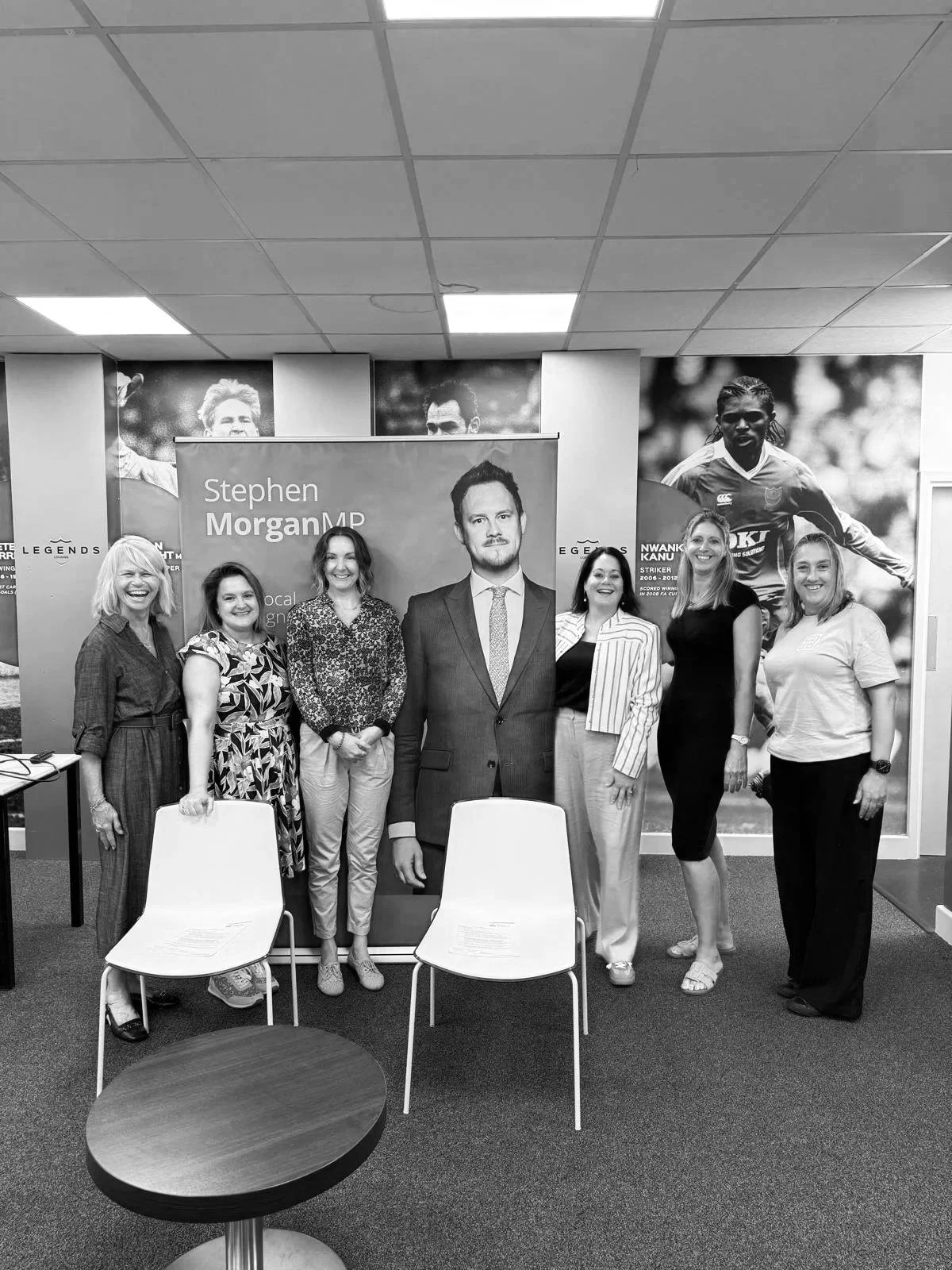 Group of seven women standing in front of a large poster of a man named Stephen Morgan MP, with two chairs and a small round table in front, in a room with sports-themed posters and a ceiling with fluorescent lights.