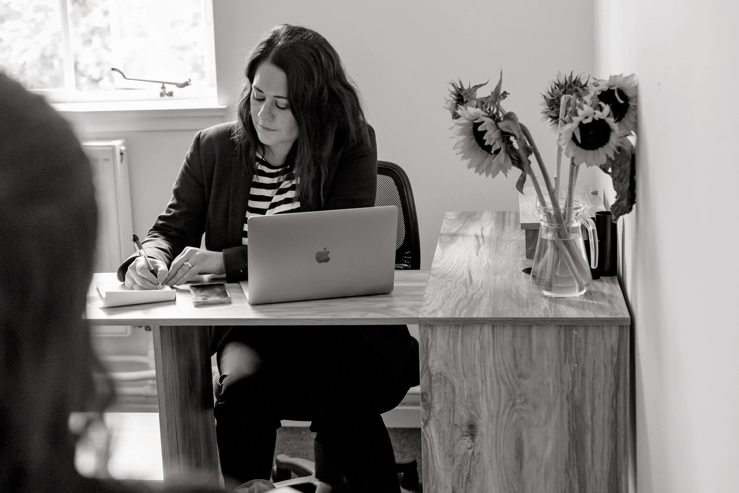 A woman sitting at a desk writing in a notepad with a MacBook in front of her, and a vase of sunflowers on the side.