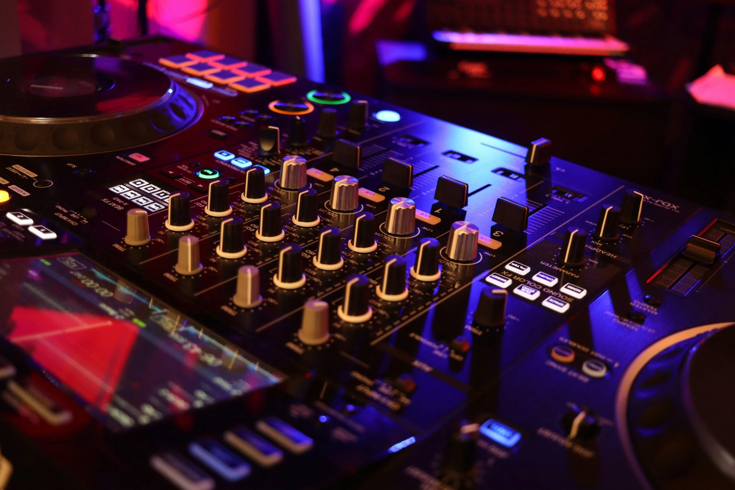 A close-up of a professional DJ controller with illuminated knobs and buttons, set in a dark, colorful lighting environment.