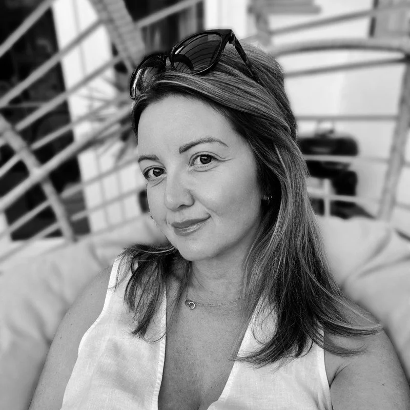 A woman with long hair wearing sunglasses on her head and a sleeveless top, sitting outdoors with blurred background.
