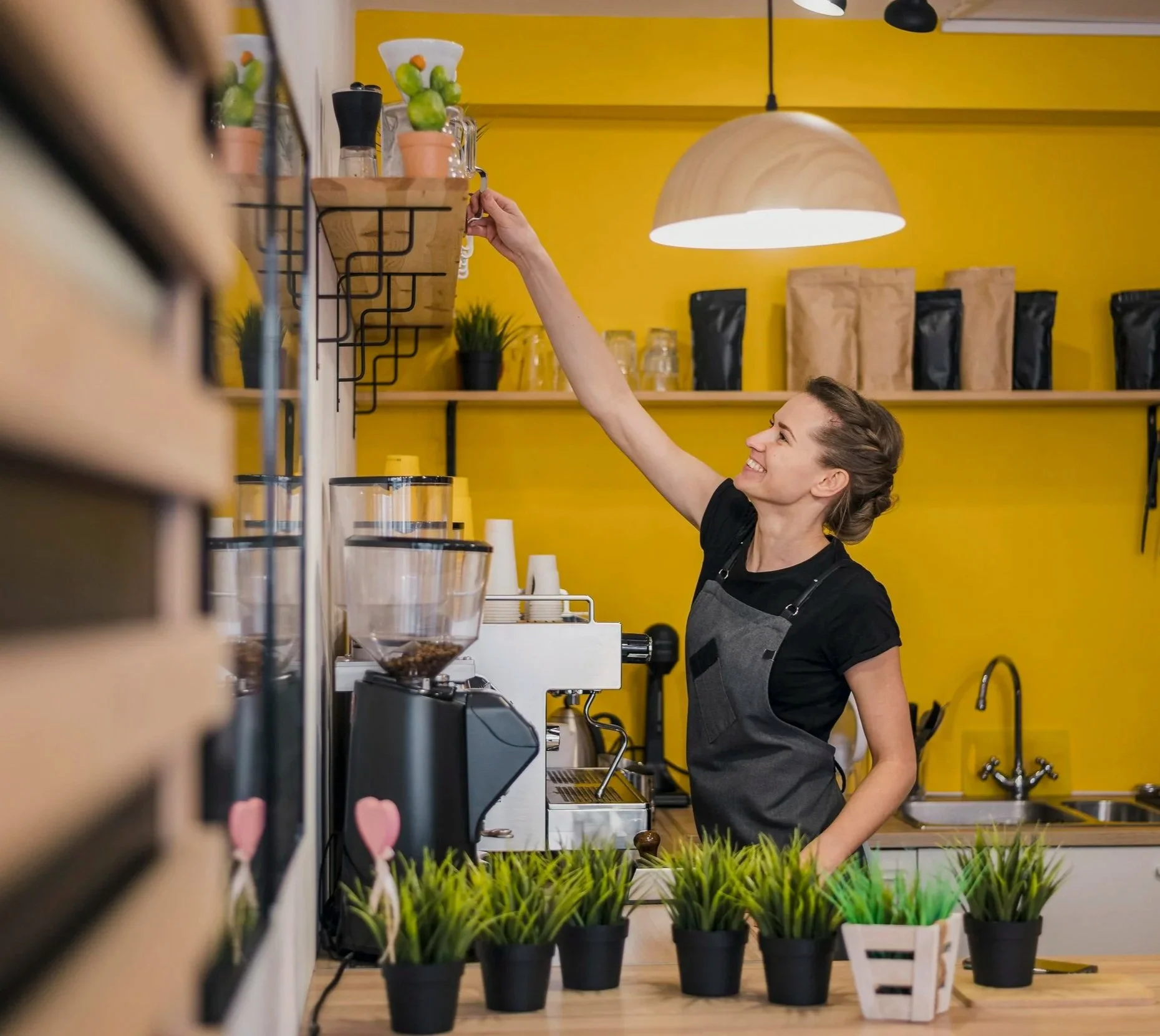 A woman working behind a coffee counter, reaching for an item on a shelf, with yellow walls and various coffee supplies and pots in the background.