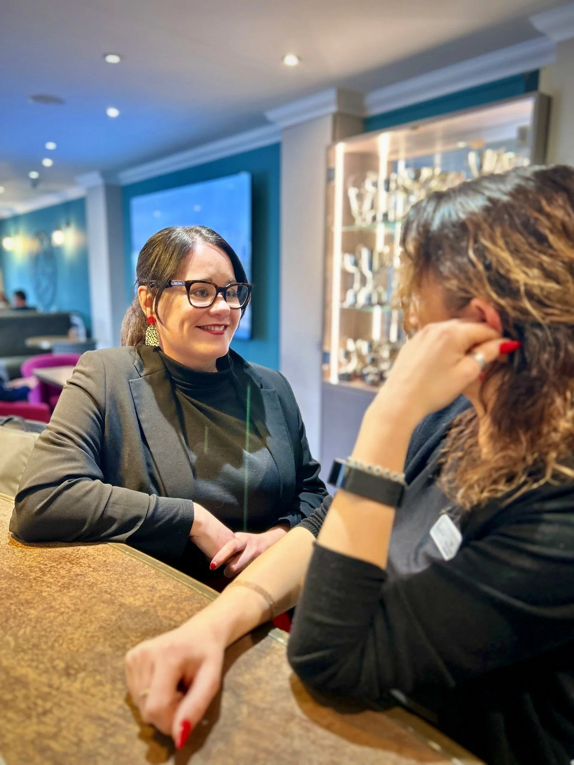 Two women in conversation at a restaurant or cafe, with one woman smiling and wearing glasses, and the other woman resting her head on her hand, wearing a watch and with curly hair.
