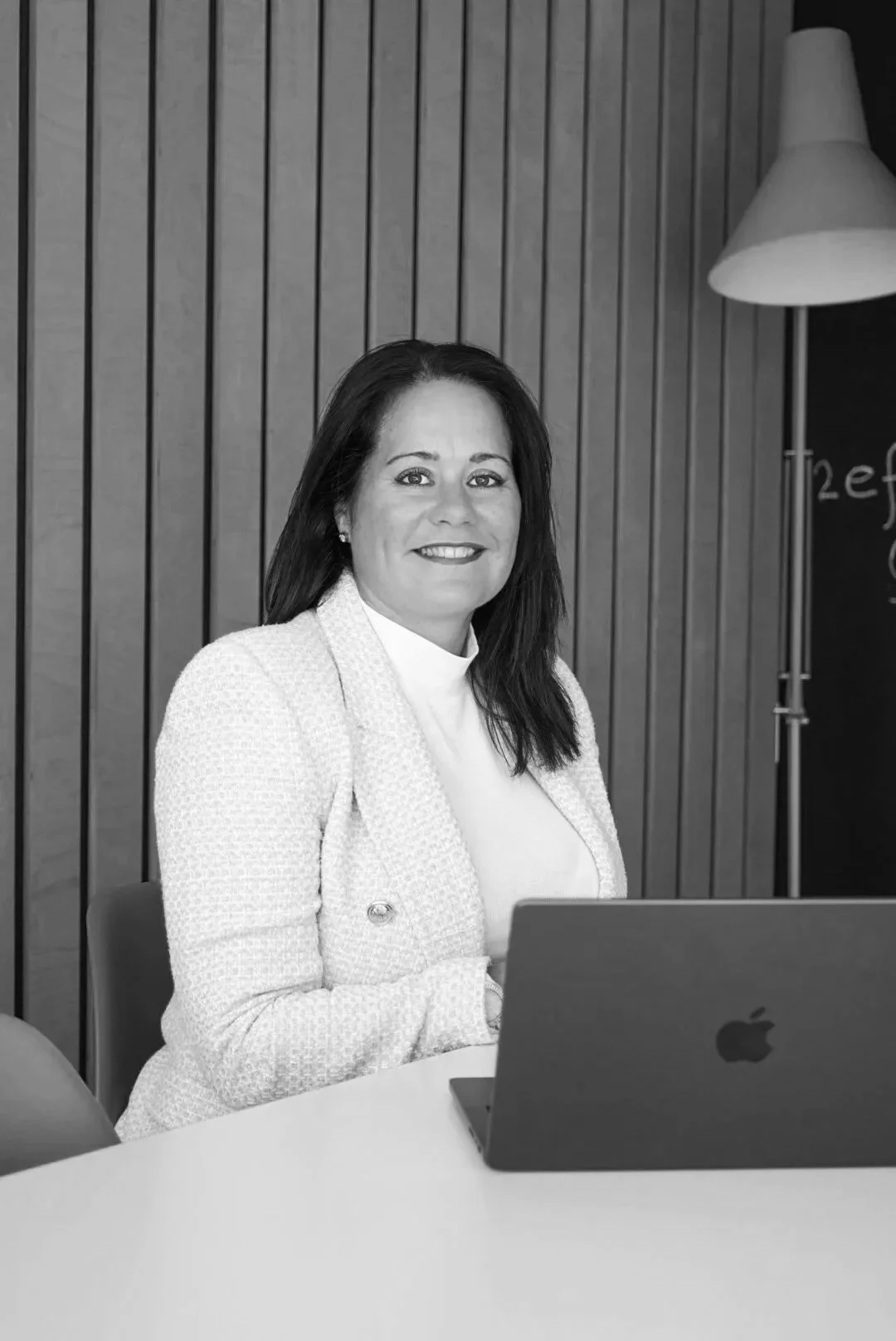 Black and white photo of a woman with dark hair smiling at the camera, seated at a desk with a laptop, in an office setting with wooden slat walls and a floor lamp in the background.