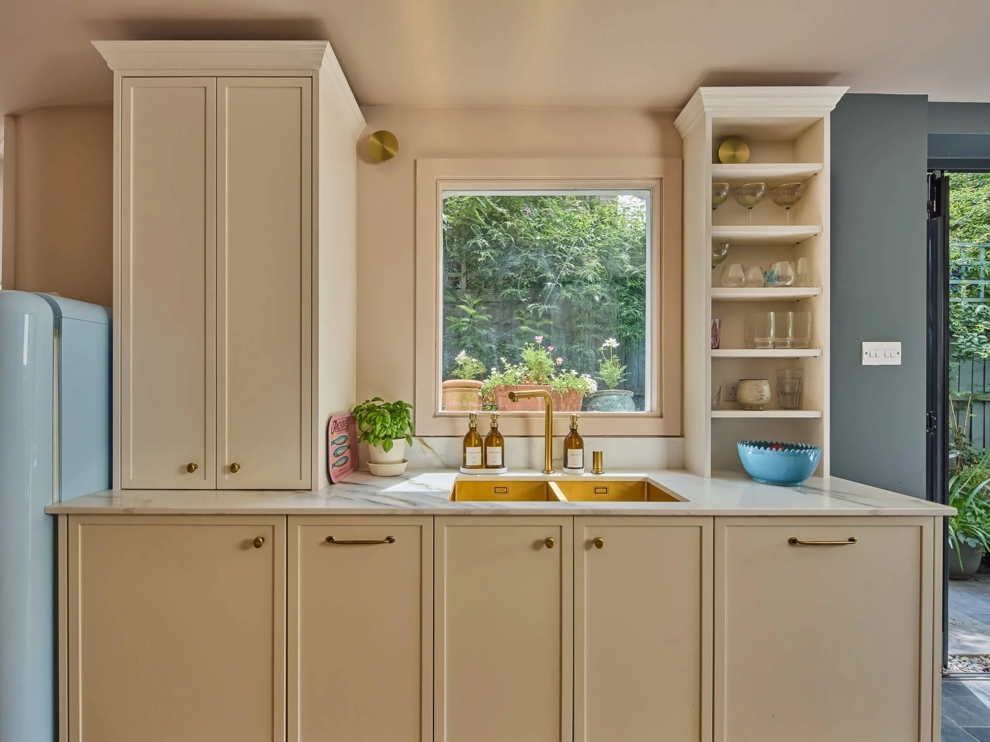 Elegant kitchen with brass sink, marble worktops, garden views, shelving.