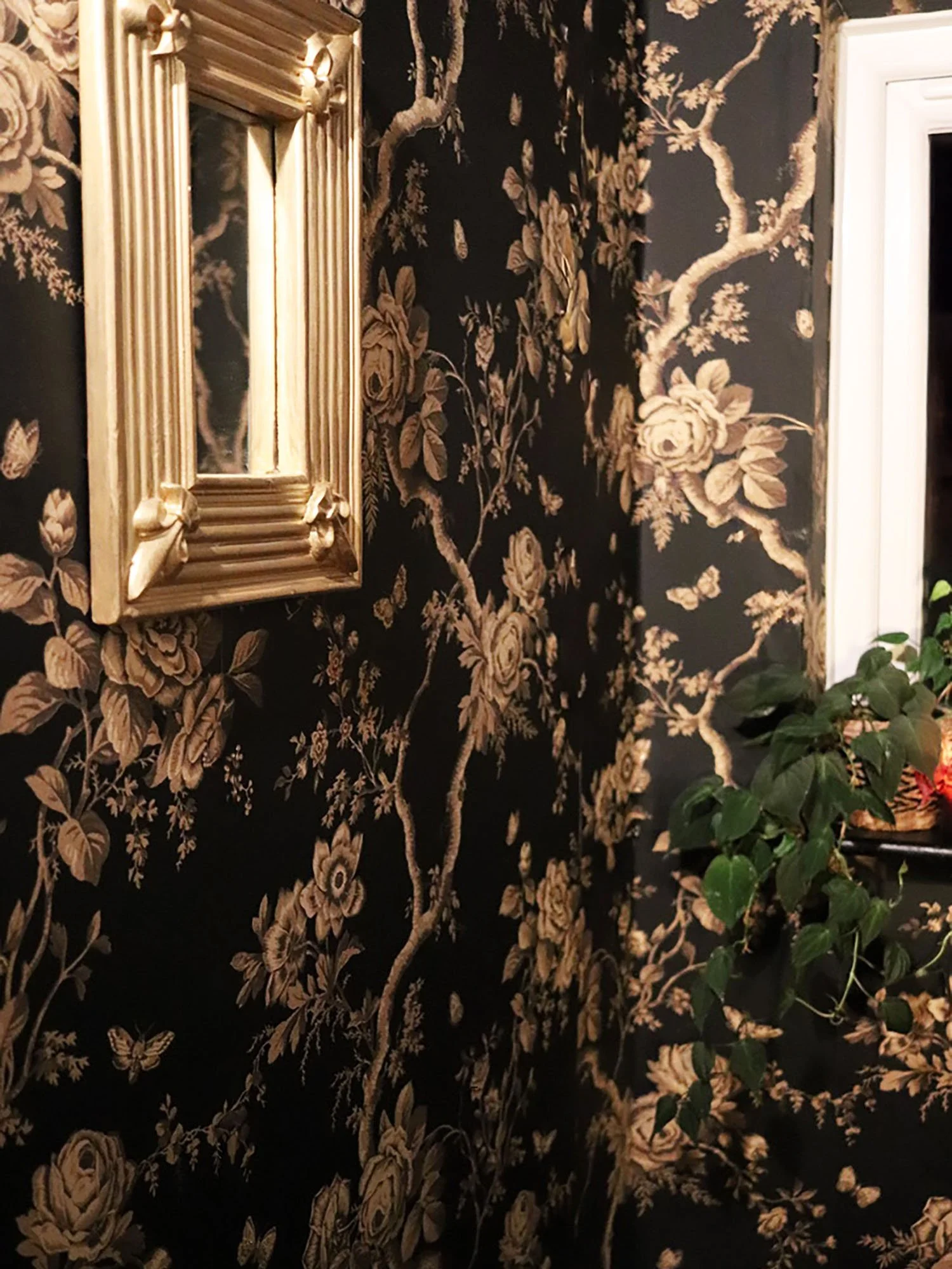Hallway featuring black-and-gold floral wallpaper with gold-framed mirror and plant