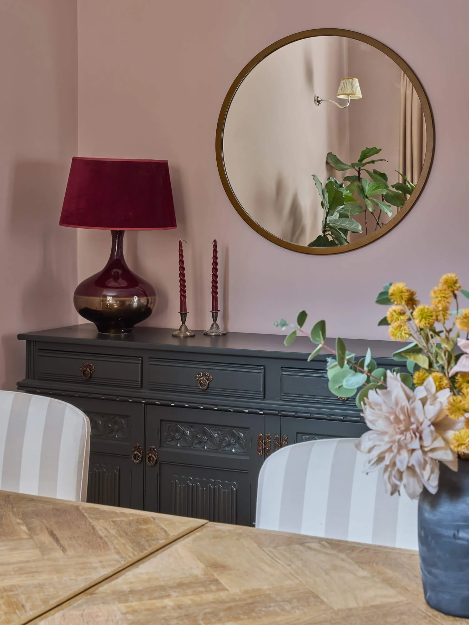 Blush dining room with painted sideboard, round mirror and floral arrangement displayed.