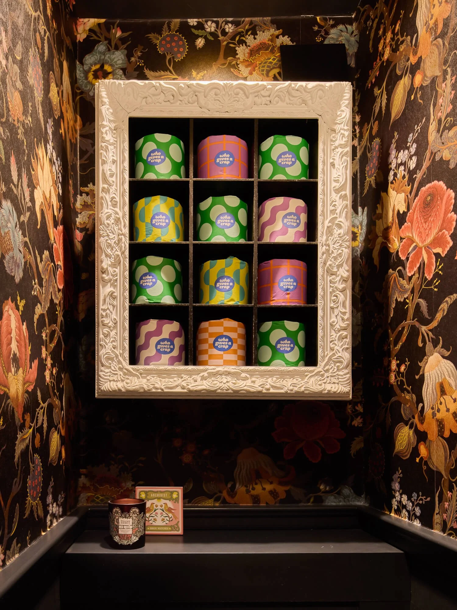 Dramatic cloakroom with floral wallpaper, framed storage, colourful toilet rolls, display.