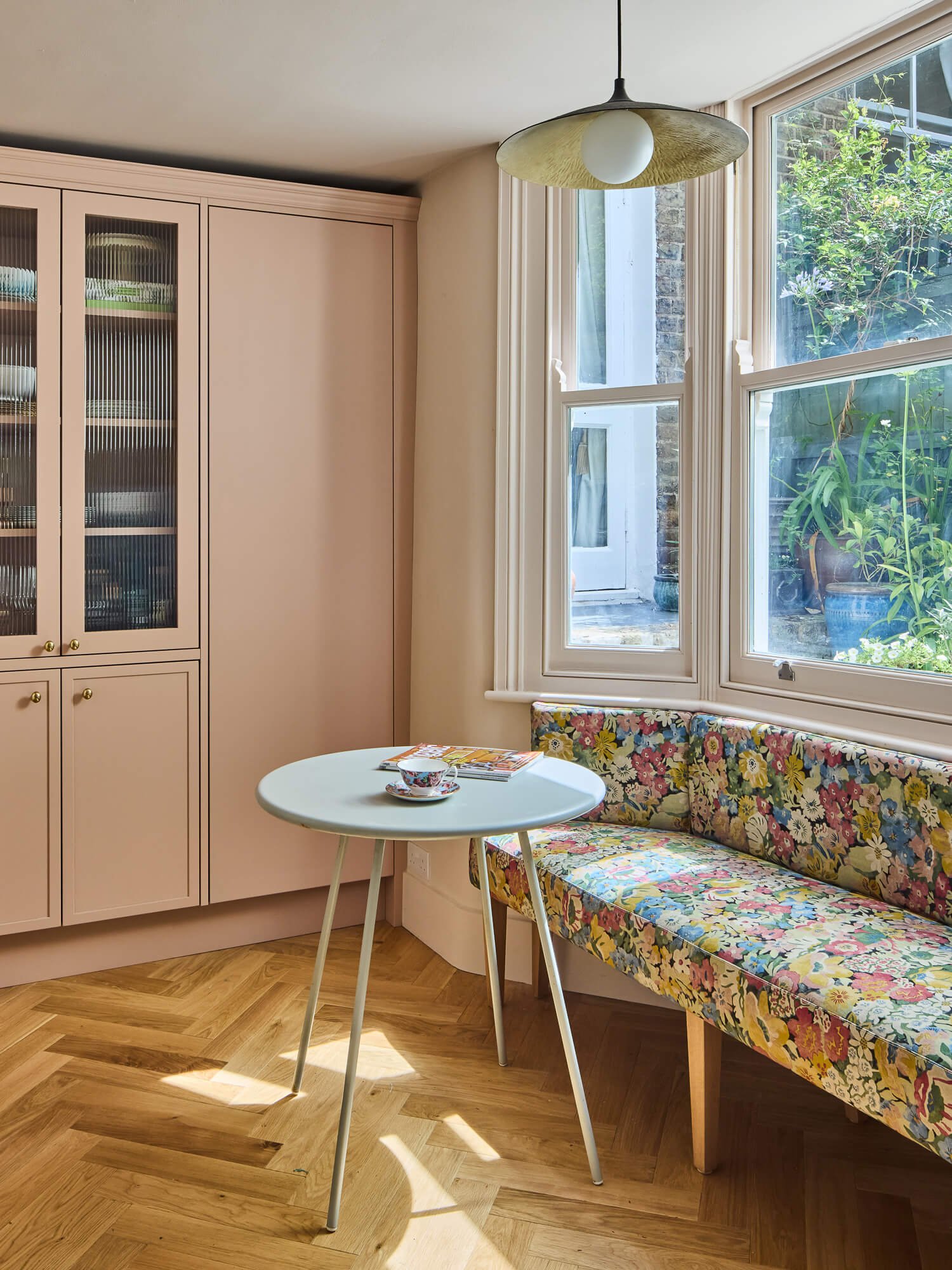 Cosy breakfast nook with blush cabinetry, floral banquette, herringbone flooring.