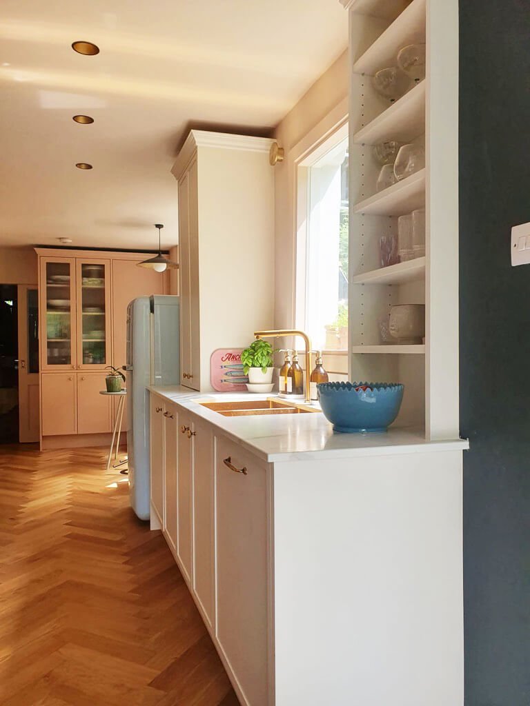 Elegant kitchen with brass sink, herringbone flooring, blush cabinetry, shelving.