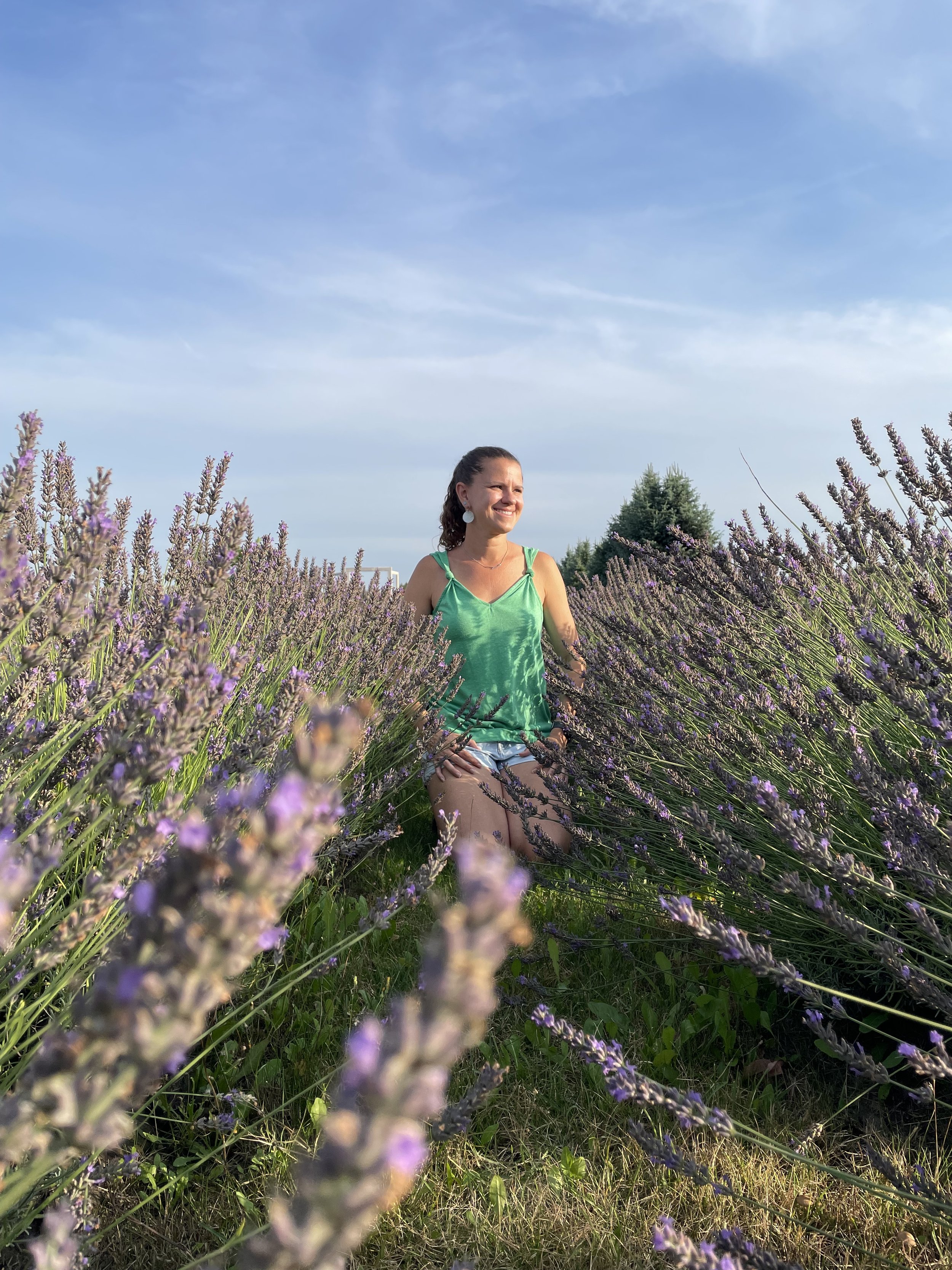 Woman kneeling among purple lavender flowers outdoors, wearing a green top and denim shorts, smiling with a blue sky and trees in the background.