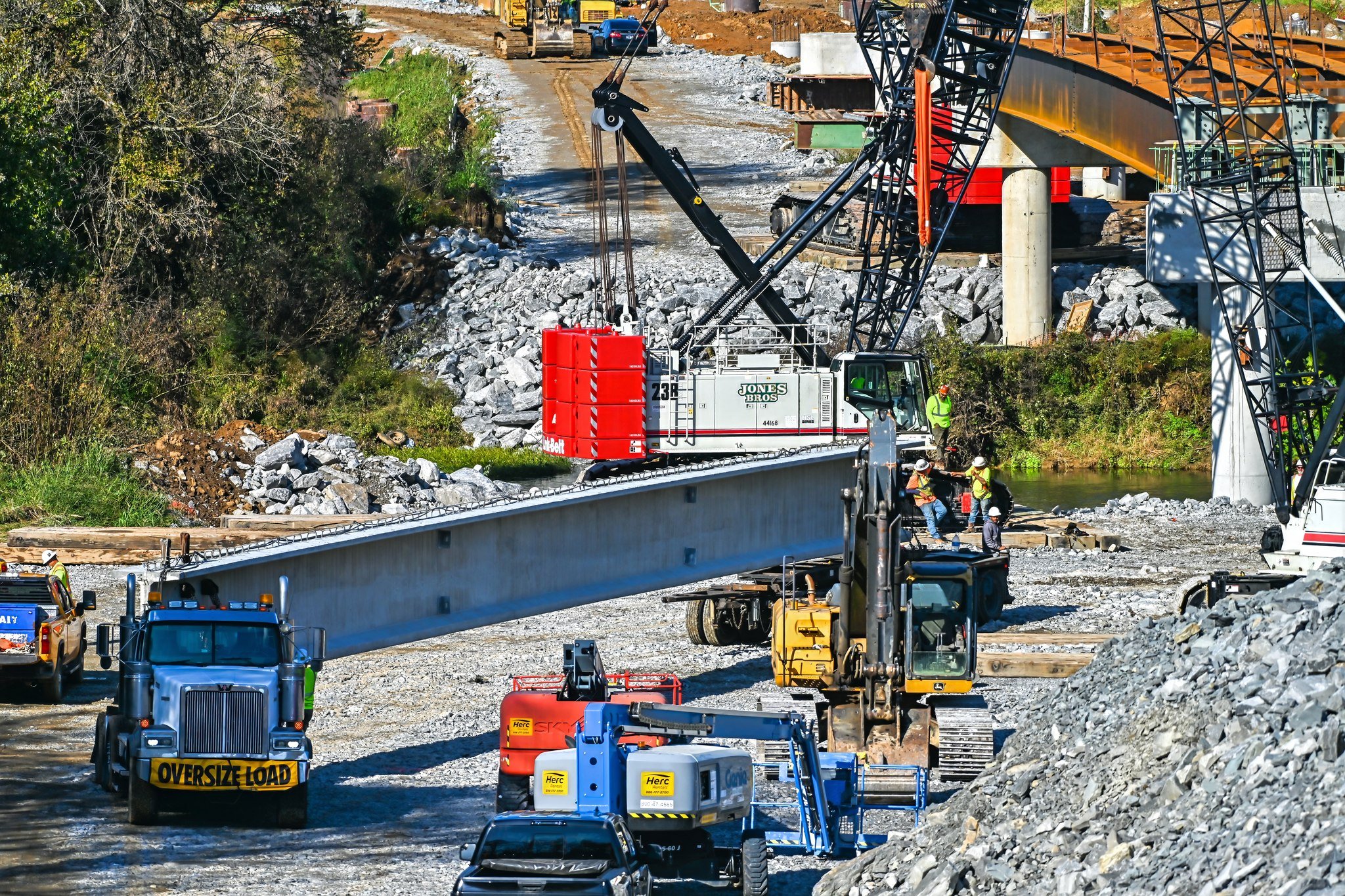 Construction workers using heavy machinery, including cranes, excavators, and trucks, working on a bridge project over a rocky landscape with dirt and gravel, with some workers wearing orange and yellow safety vests and helmets.