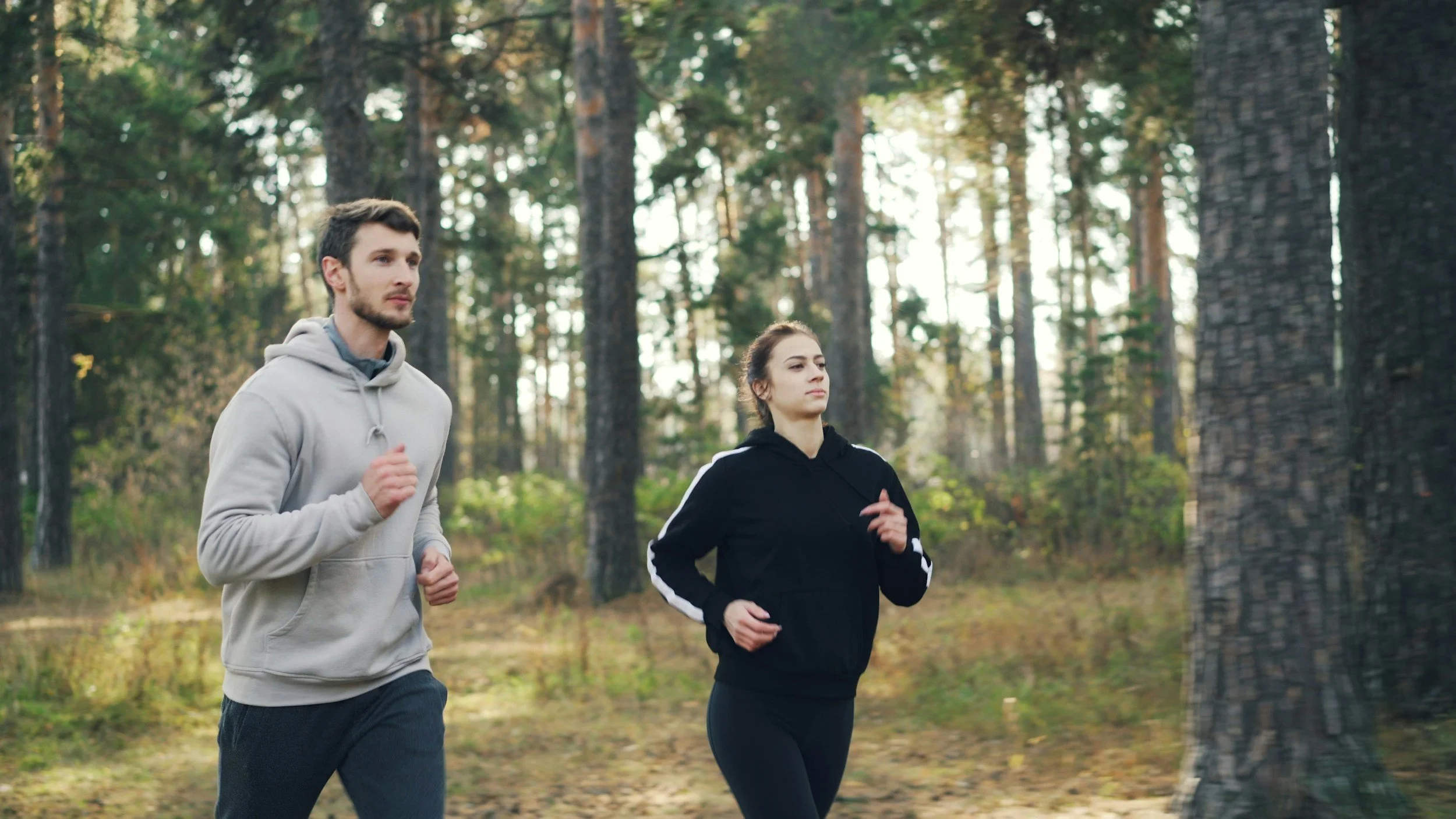 A man and woman jogging in a forest during daytime, surrounded by tall trees and natural scenery.