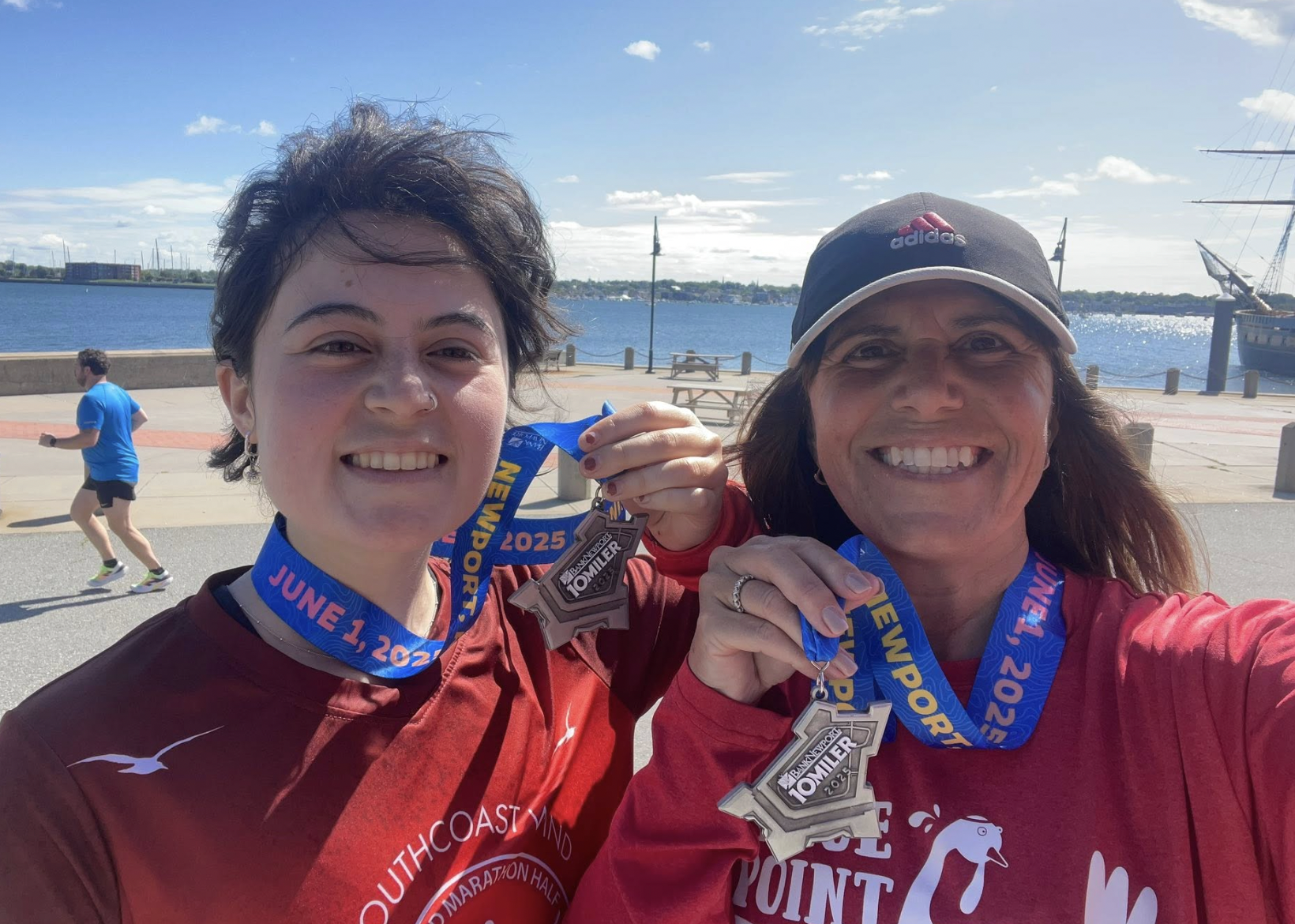 Two women with medals around their necks taking a selfie outdoors by a body of water, with a jogger in the background and ships visible in the distance.