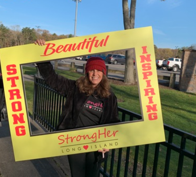 A woman smiling and holding a large yellow frame that says 'Beautiful,' 'Strong,' 'Inspiring,' and 'Strong Her Long Island.' She is outdoors near a fence and cars, wearing a red knit hat and a dark jacket.