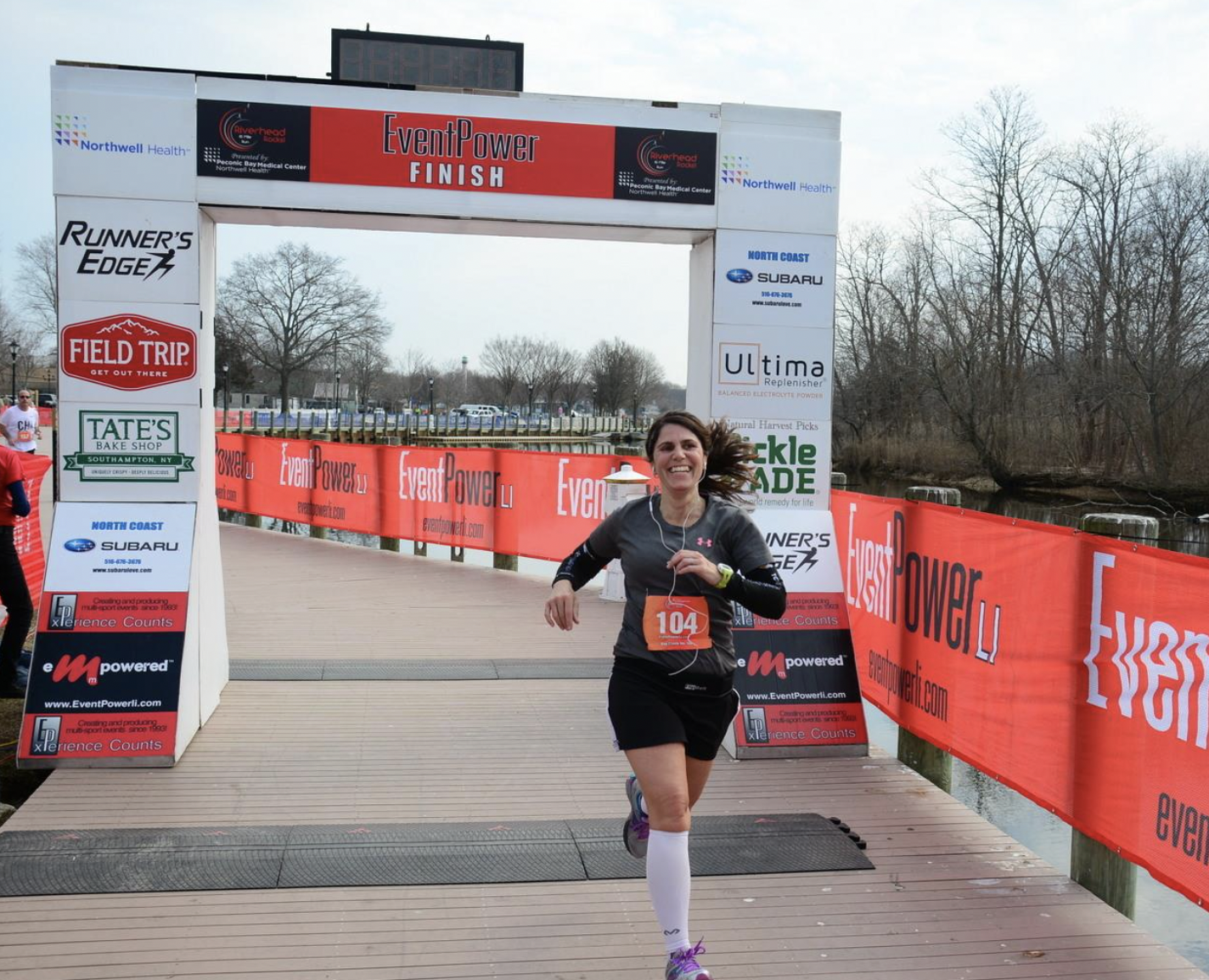 A woman crossing the finish line of a race with a smile, wearing a black athletic outfit, white compression socks, and a race bib number 104, during an outdoor event on a cloudy day.