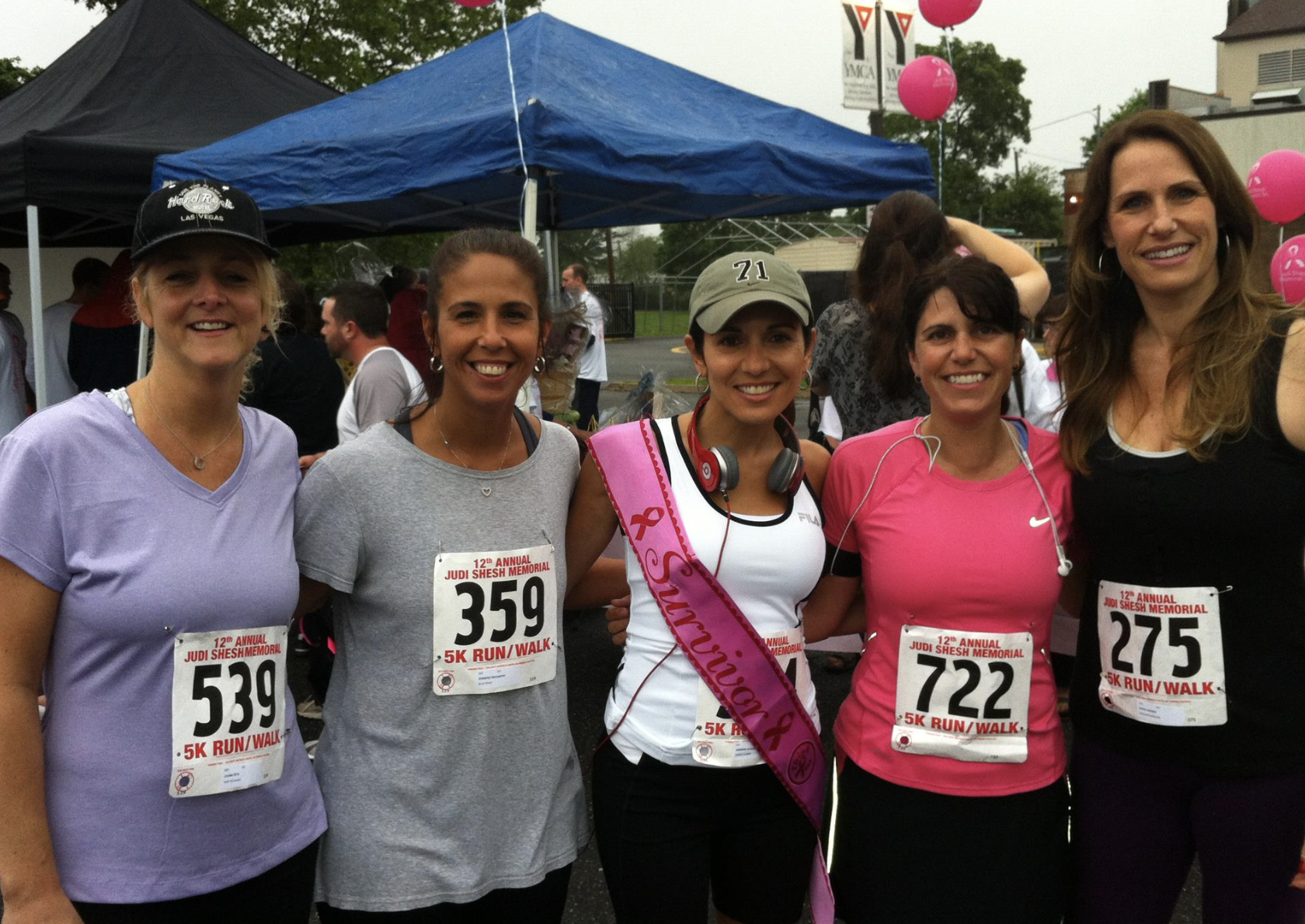 Group of five women standing outdoors at a charity run event, wearing race bibs and sports attire, smiling for a photo, with tents and banners in the background.