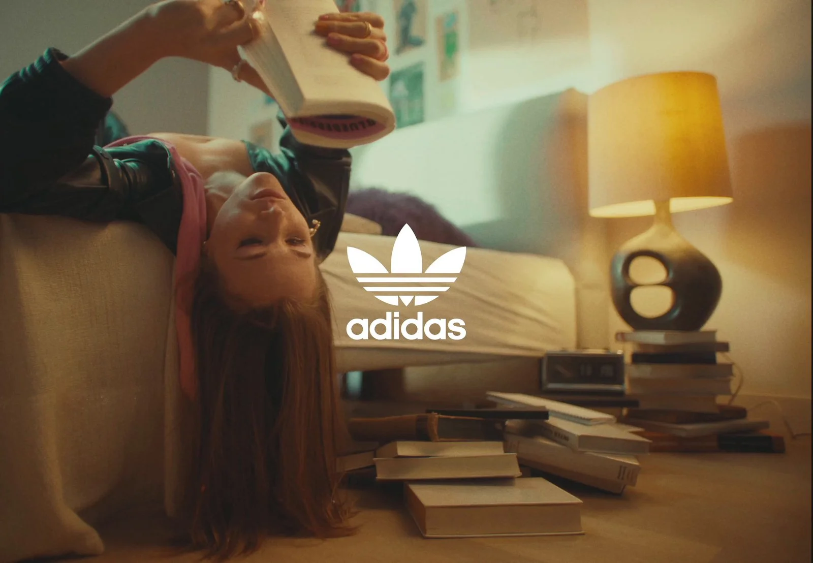 A young girl with red hair hangs upside down from a sofa, reading a book. There are multiple books on the floor and a stack on a side table. A lamp on a bedside table provides warm lighting, and a wall with pictures is in the background. The Adidas l