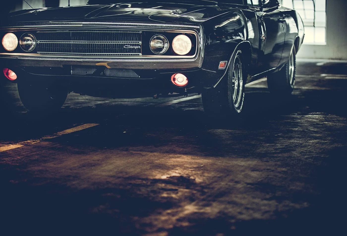 A vintage black Dodge Charger muscle car parked in a dimly lit garage with concrete floor and windows in the background.