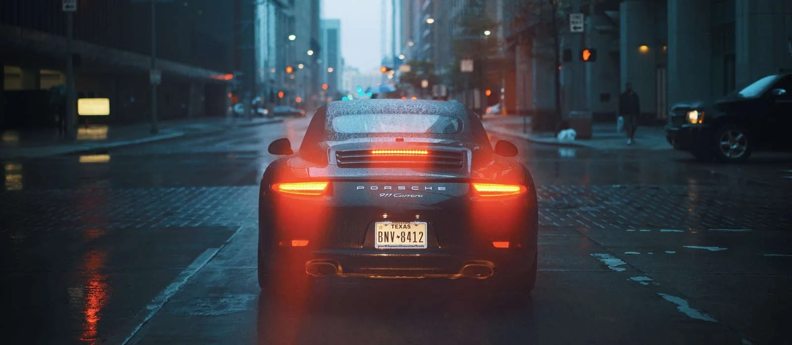 Rear view of a black Porsche 911 Carrera with illuminated taillights, driving on a city street in rainy weather during dusk or night.