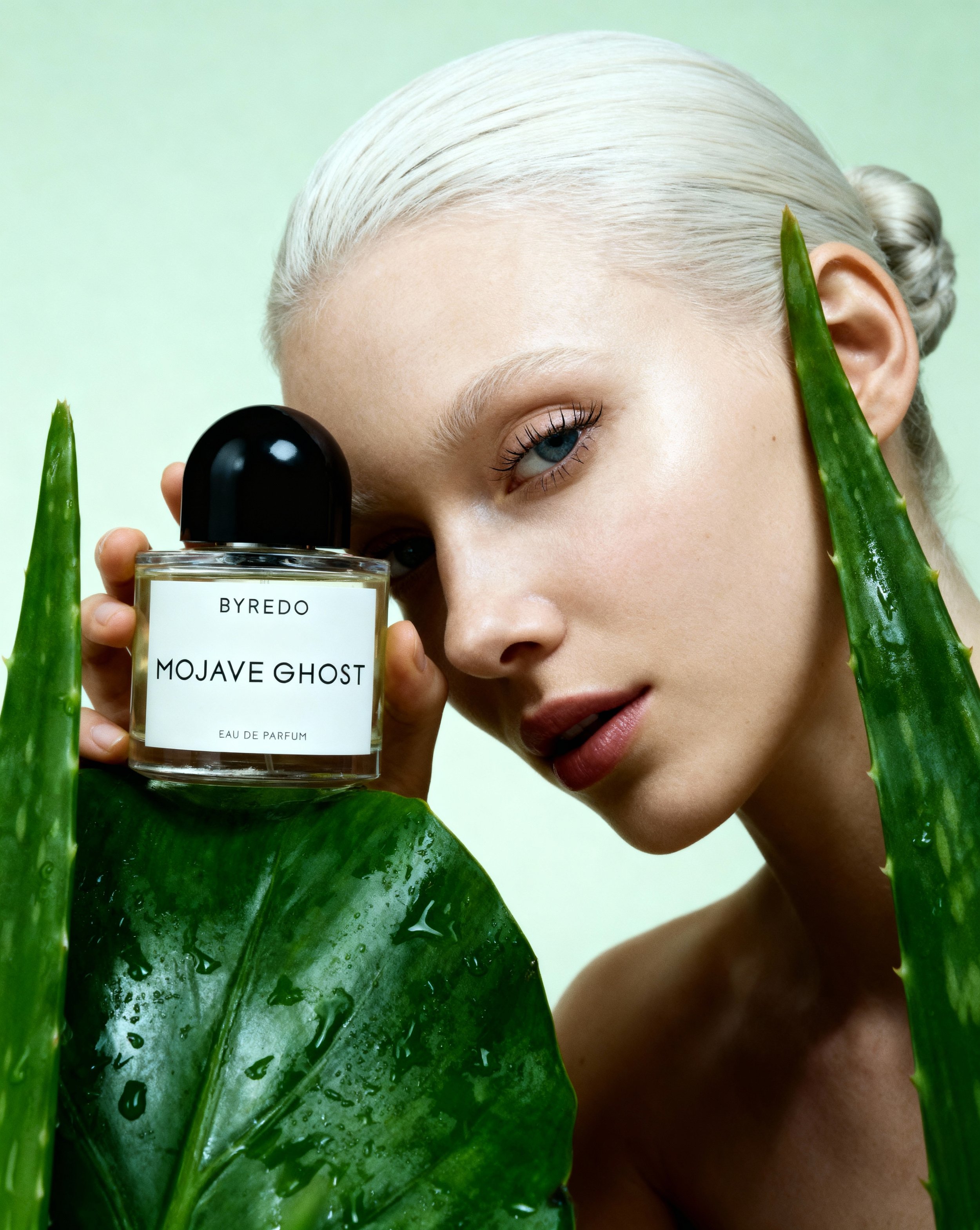 A woman with platinum blonde hair holds a perfume bottle labeled 'Byredo Mojave Ghost' close to her face, surrounded by large green aloe vera leaves.