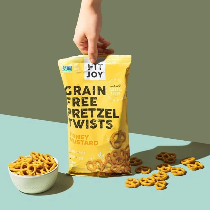A hand holding a yellow bag of Fit Joy Grain Free Pretzel Twists with honey mustard flavor, with some pretzels spilled on a green and blue surface, and a bowl of pretzels next to it.