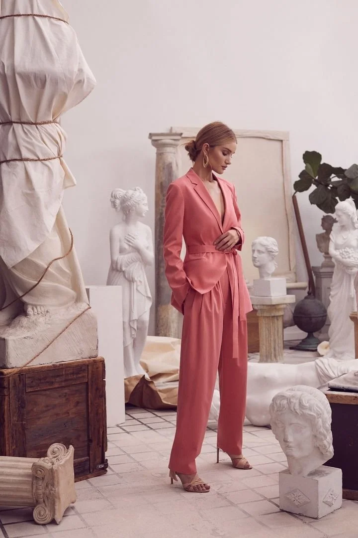 A woman in a pink pantsuit and high heels stands with her hands on her stomach in a room filled with classical sculptures and statues.