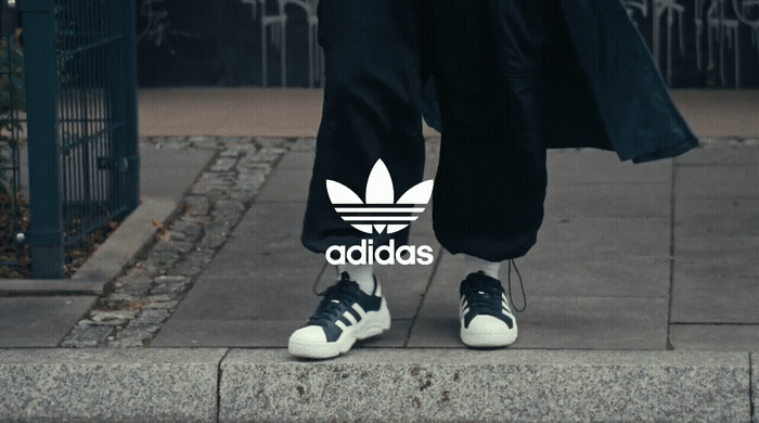 Person standing on a sidewalk wearing black pants and Adidas sneakers with the Adidas logo overlayed in the center of the image.