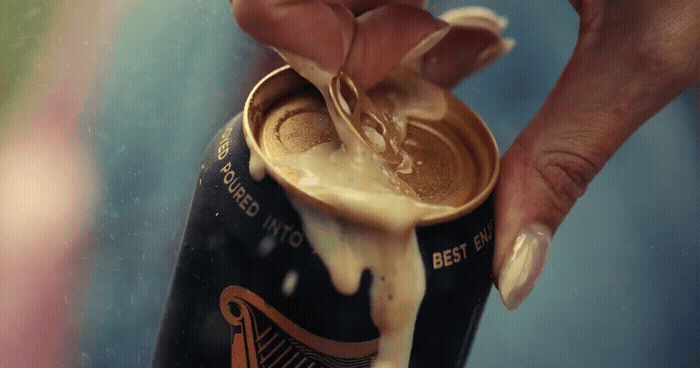 A hand opening a beer can with foam spilling out.