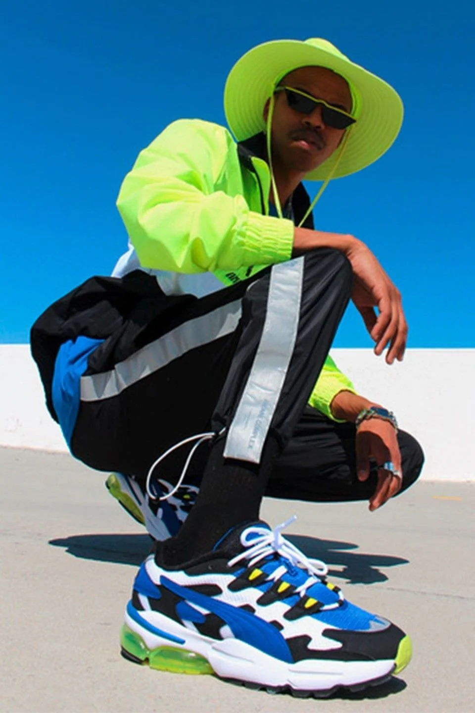 A person dressed in neon yellow and black athletic clothing, wearing a large wide-brimmed neon yellow hat, sunglasses, and crouching on a rooftop with a blue sky background.