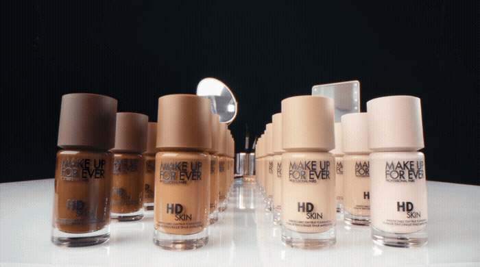 Multiple bottles of Make Up For Ever HD Skin foundation arranged in a row on a reflective surface, with a mirror in the background.