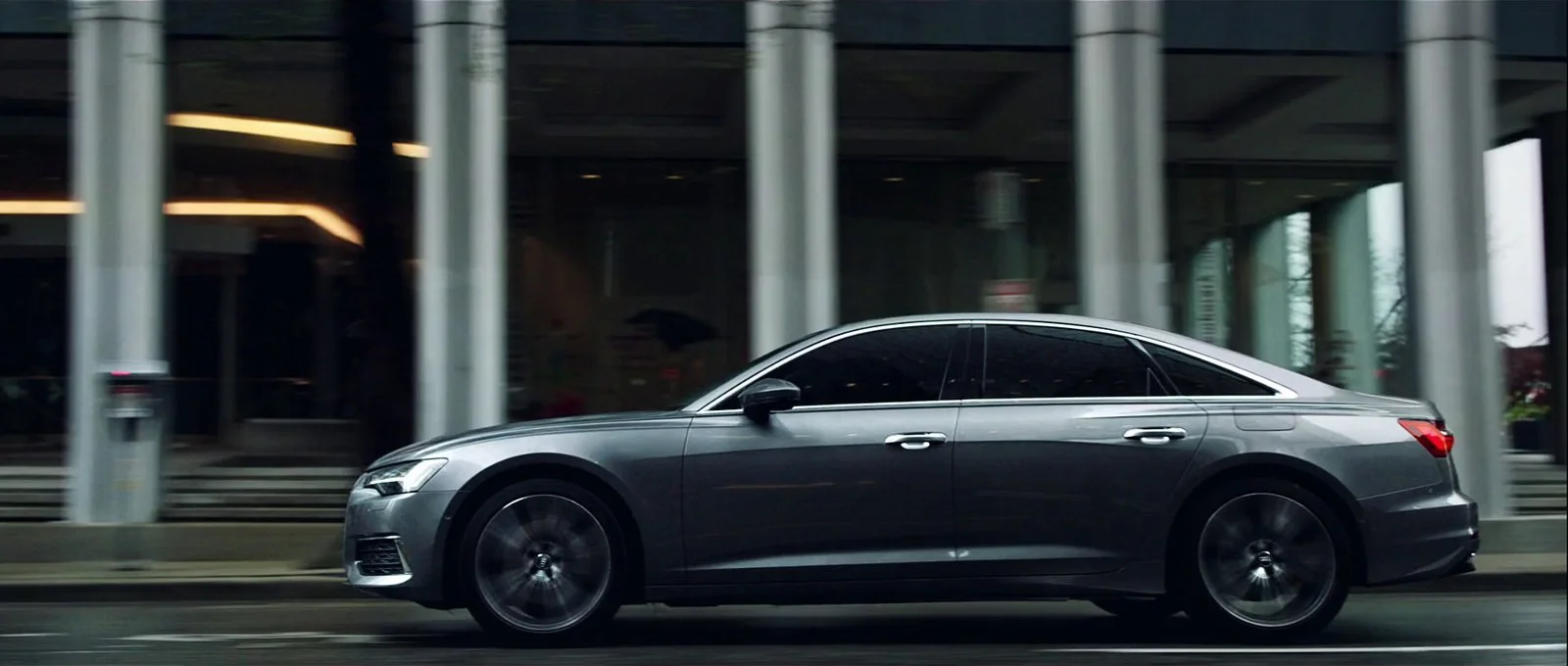 A sleek gray luxury sedan driving on a city street with glass buildings in the background.